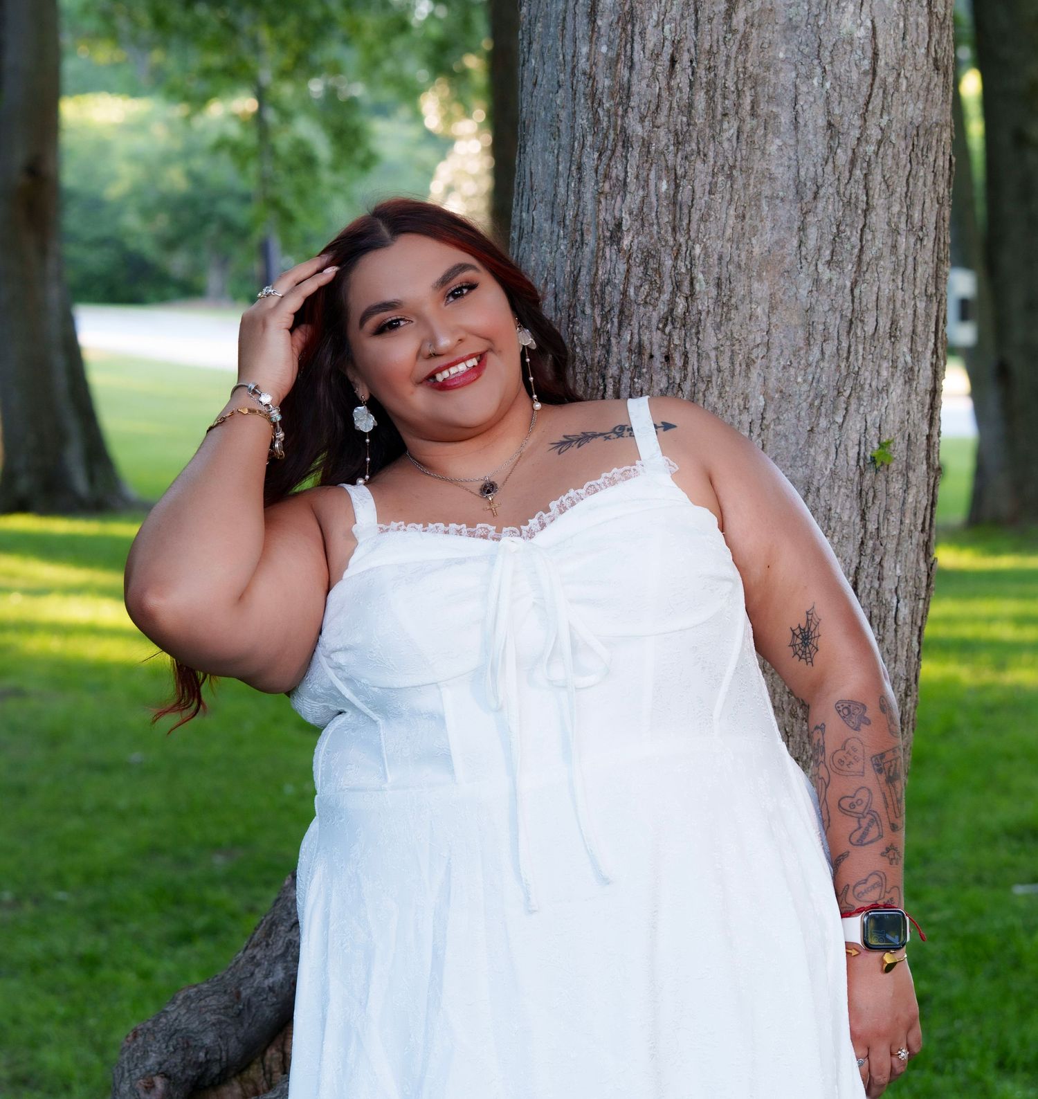 Woman in white sundress smiling and leaning against a tree in a lush green park, showcasing tattoos and elegant earrings.