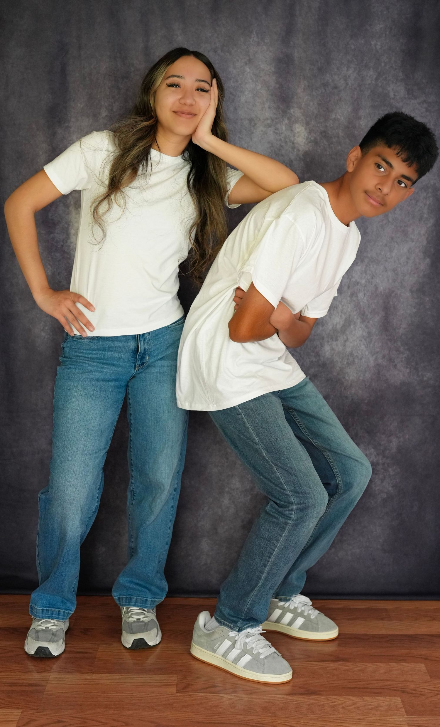 Two teens in matching white tees and blue jeans pose back-to-back in a studio, smiling confidently.