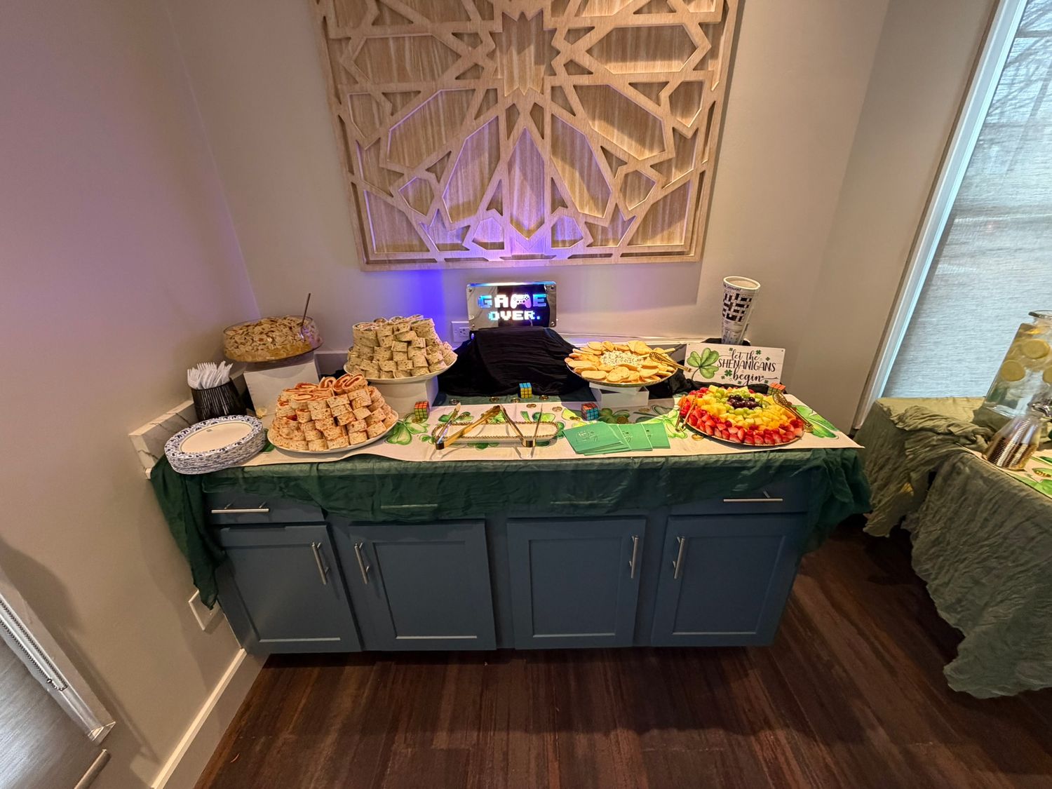 Buffet table with green tablecloth displaying assorted party foods and snacks beneath decorative wall art.