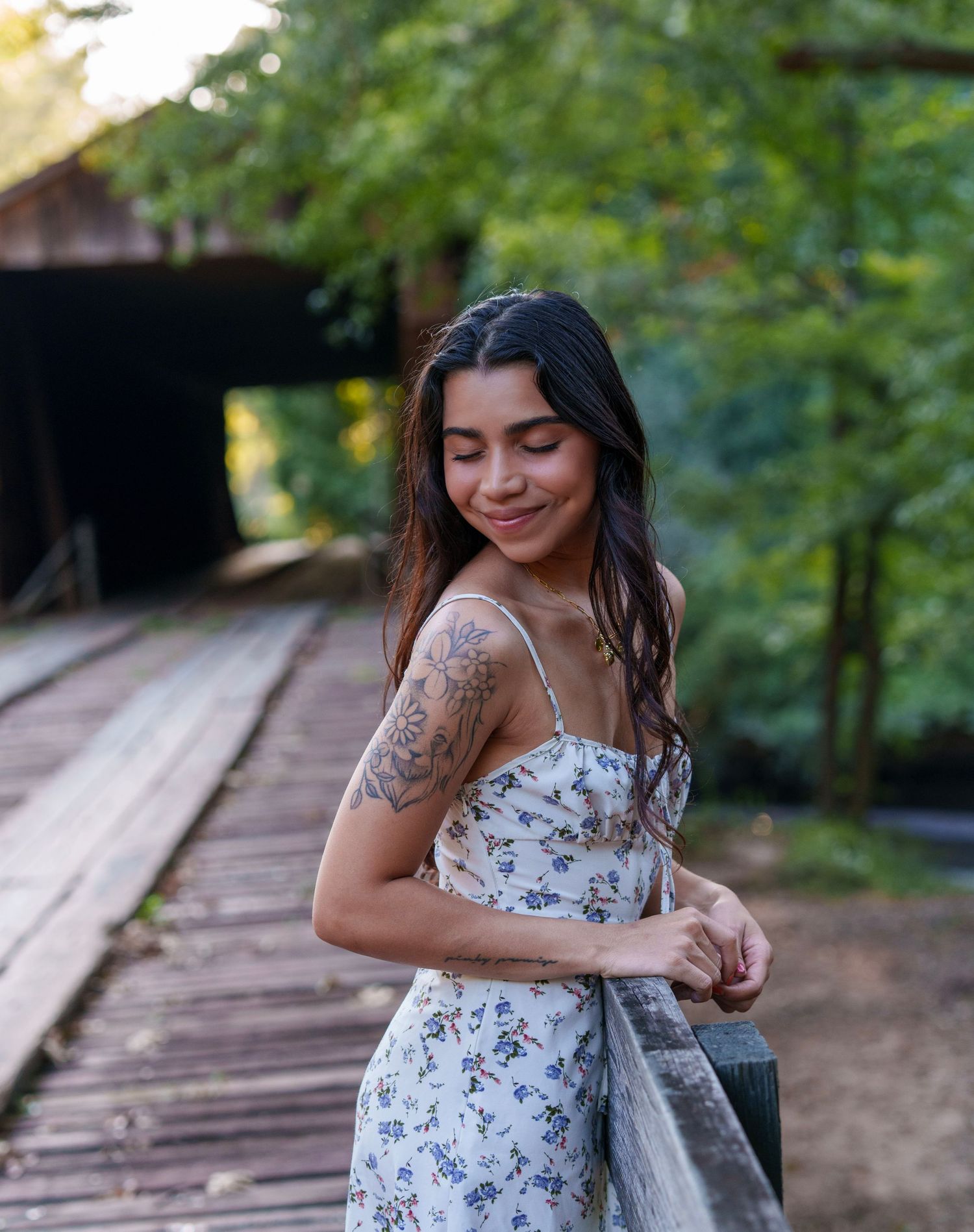 Young woman with tattoo in floral sundress smiling outdoors near wooden bridge surrounded by lush green trees.