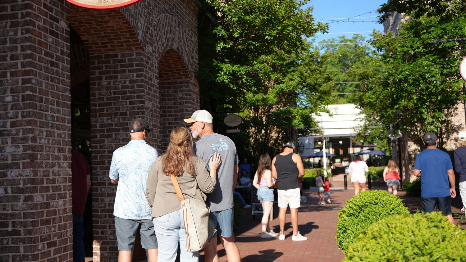 People walking along a brick-lined outdoor shopping area surrounded by lush green trees on a sunny day.