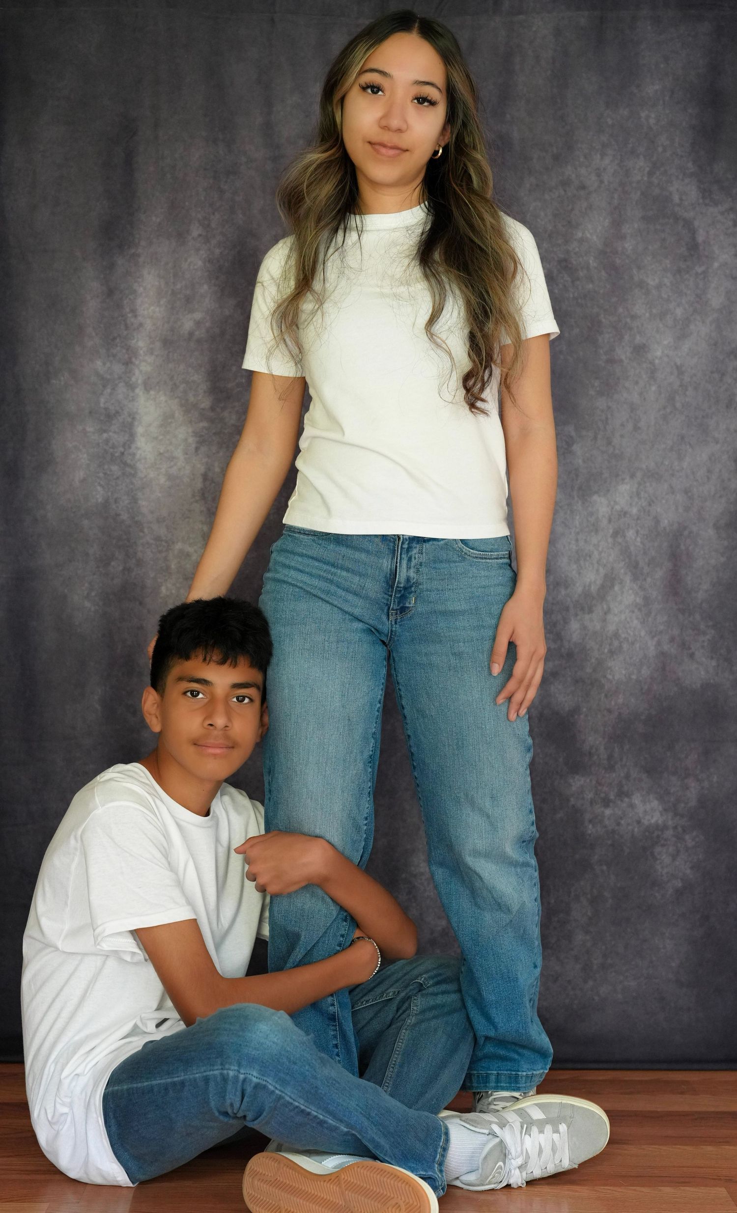 Teen girl in white tee and jeans stands behind younger boy sitting on floor in matching casual outfits.