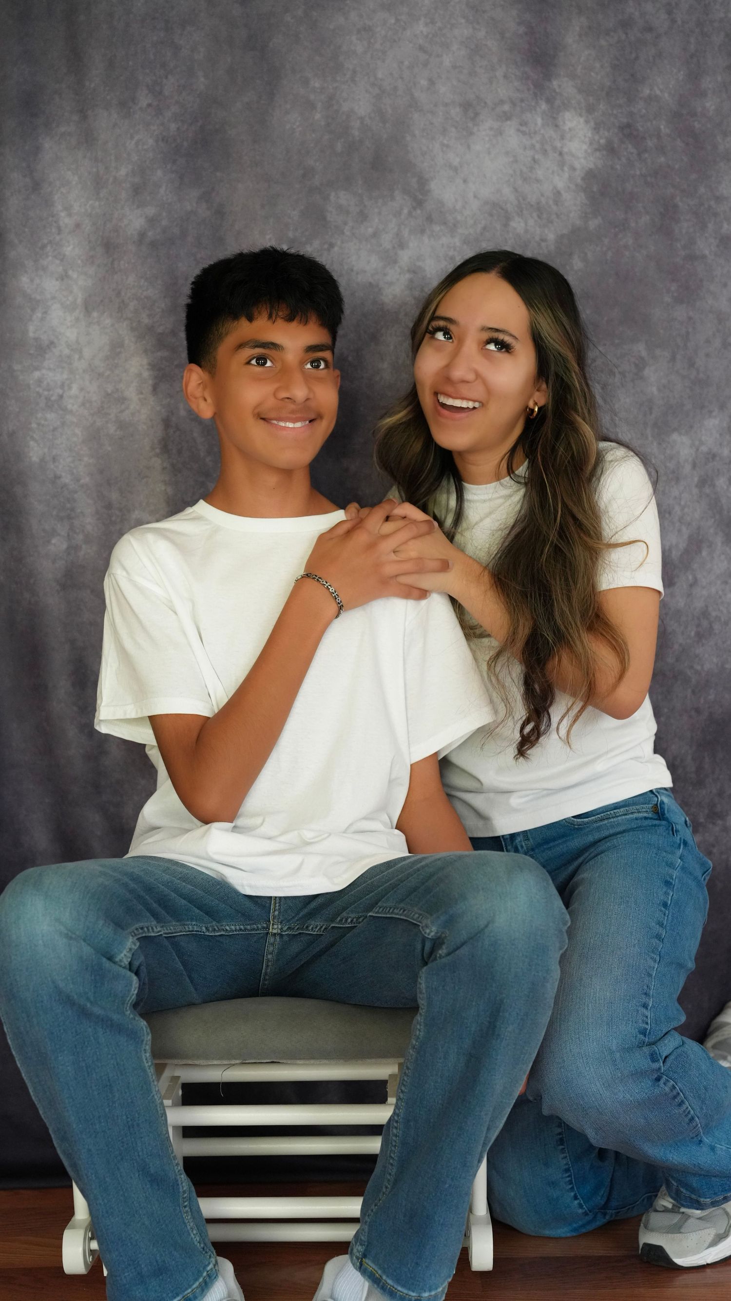 Two people in matching white t-shirts and jeans pose together smiling in front of a gray studio backdrop.