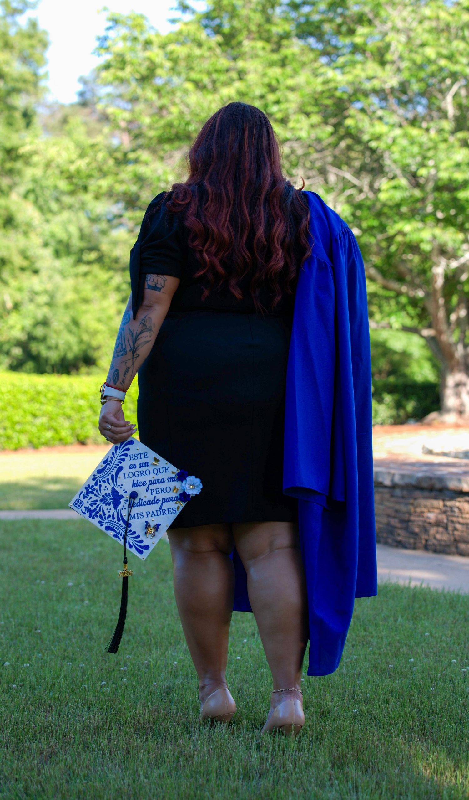 Woman in black dress holding decorated graduation cap and blue academic gown, standing on grass with trees behind.