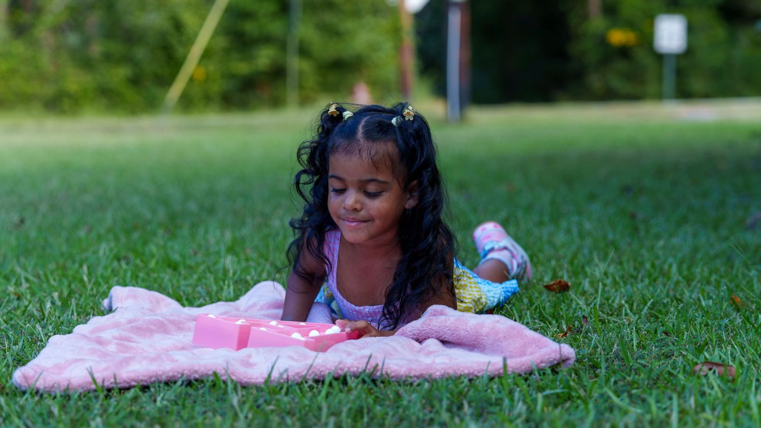 Young girl lying on pink blanket in green grass, playing with toys outdoors in a park setting.