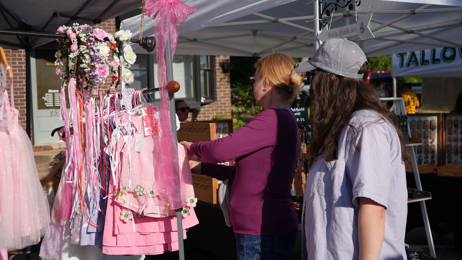 Two women browse pink dresses and floral accessories at an outdoor market vendor booth on a sunny day.