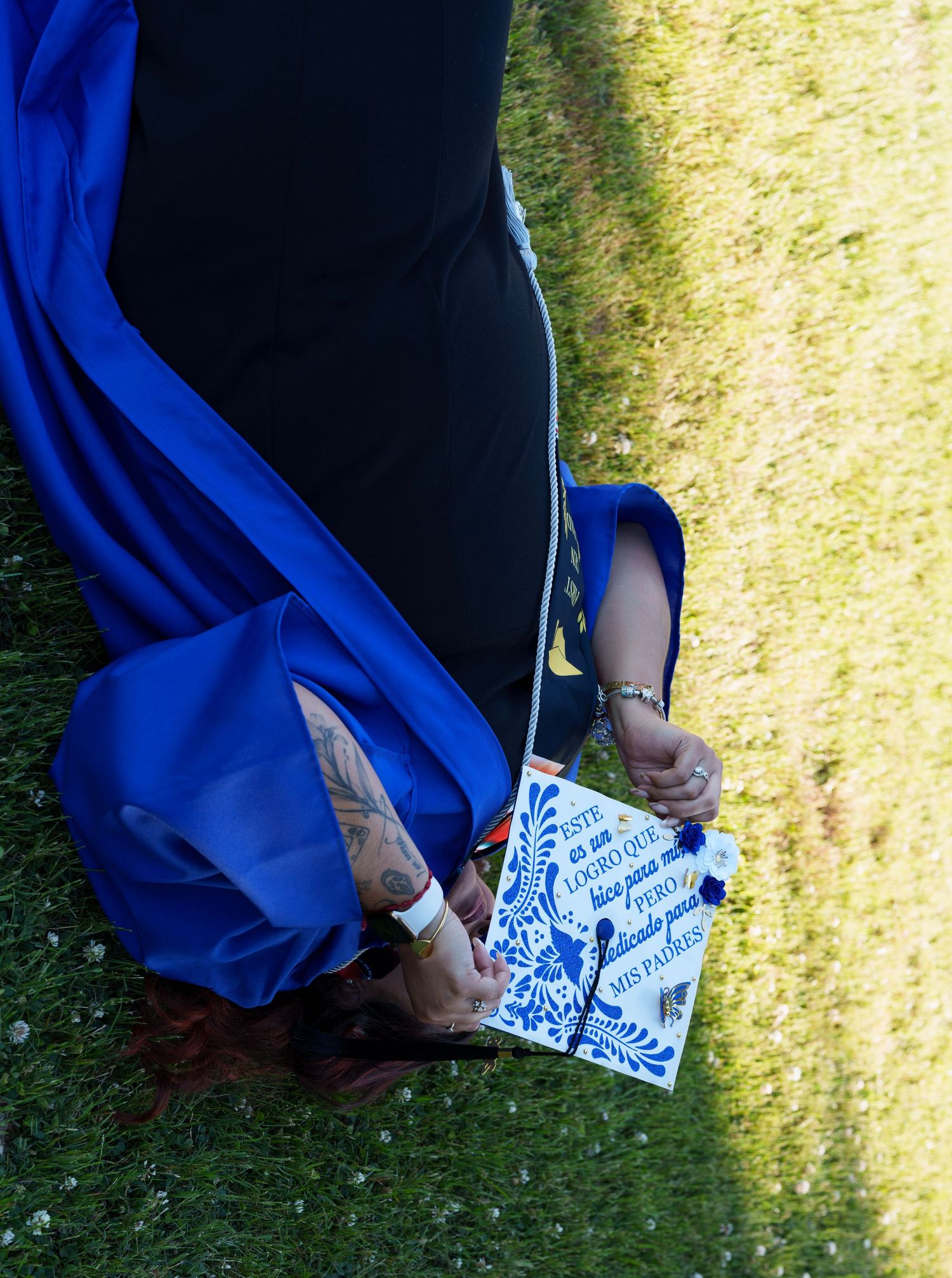 Graduate in blue cap and gown bending down on grass, holding decorated graduation cap with floral design.