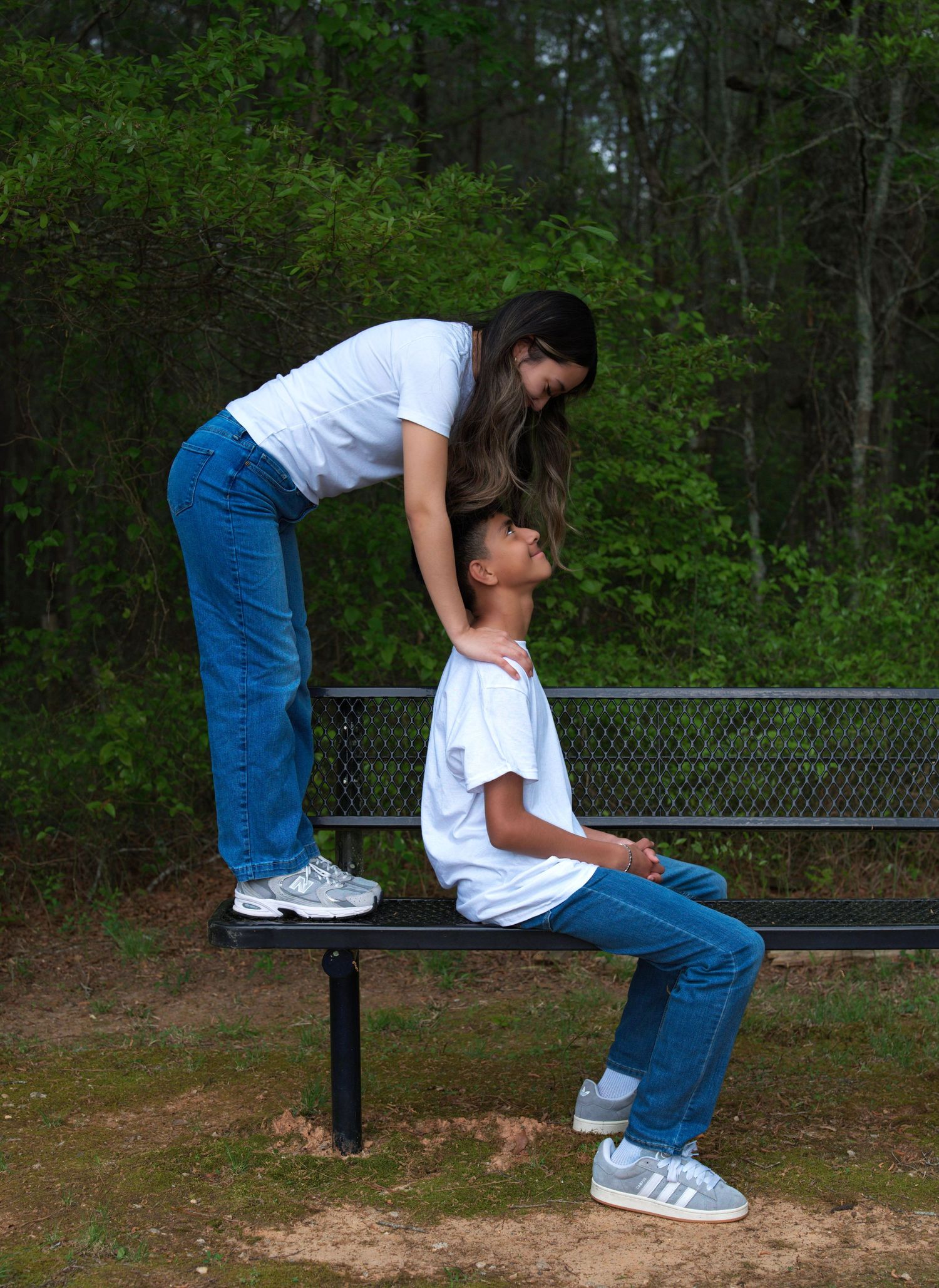 A woman standing on a bench leans over a child sitting on it, both wearing white shirts and jeans outdoors.