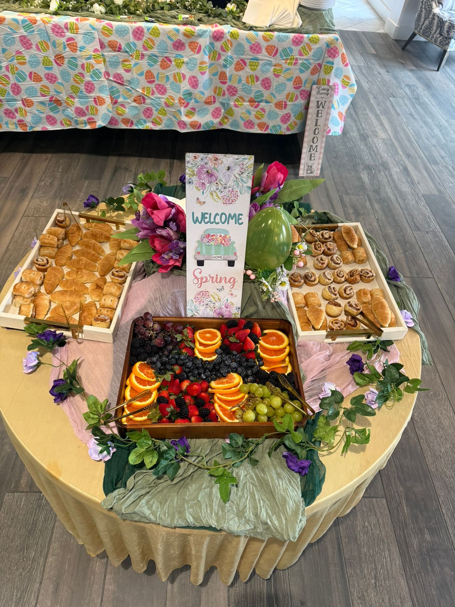 Spring-themed welcome table with fruit charcuterie board, pastries, and floral decorations at a seasonal event.