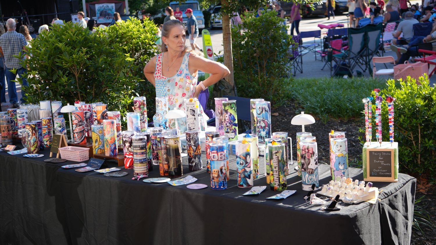 Woman standing at outdoor vendor table displaying colorful candles and gifts at a festive market event.