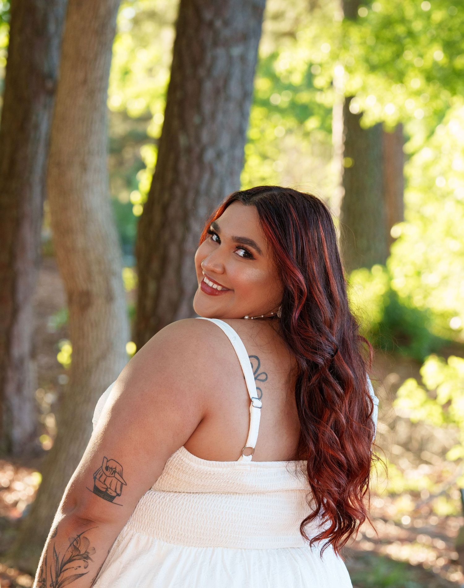 A woman with long red hair and tattoos smiles over her shoulder in a white sundress among trees.