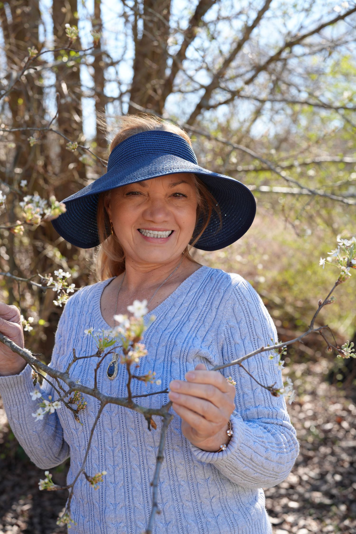 Someone wearing a blue sweater and navy sun hat stands smiling among blooming spring tree branches.