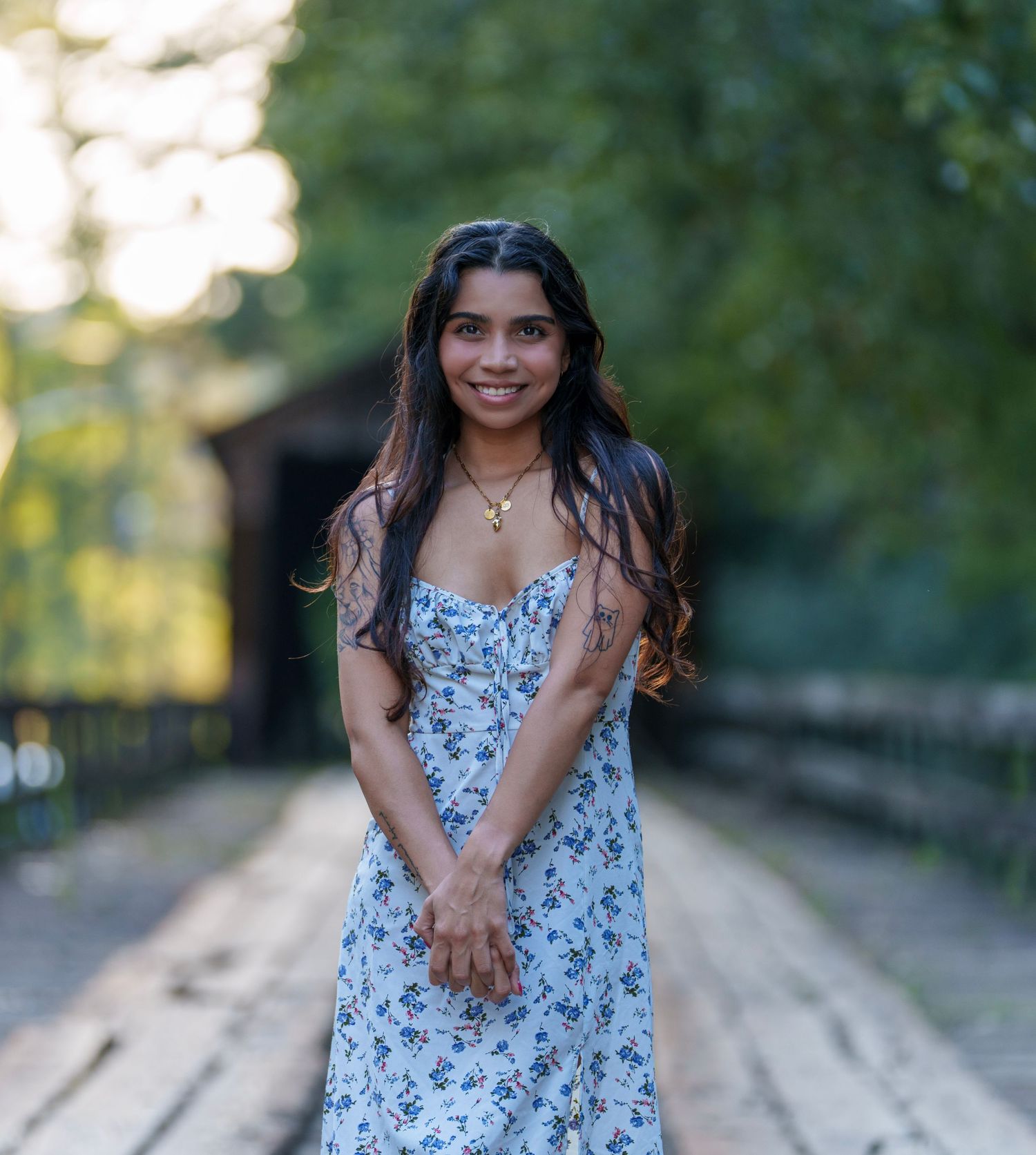 Young woman in floral sundress smiling on a wooden bridge surrounded by lush green trees.