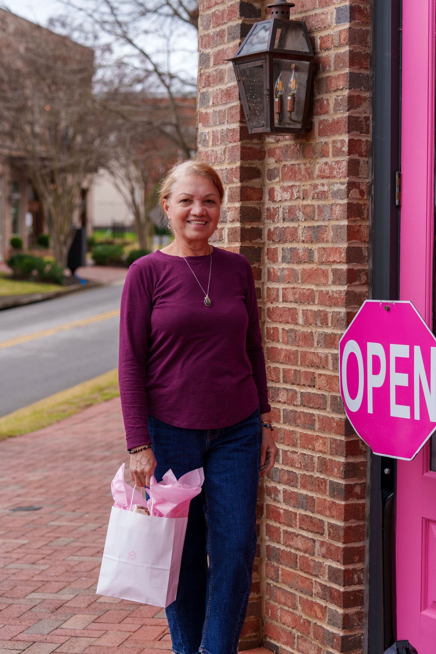 Someone in a purple sweater stands by a brick wall with a shopping bag near a bright pink open sign.