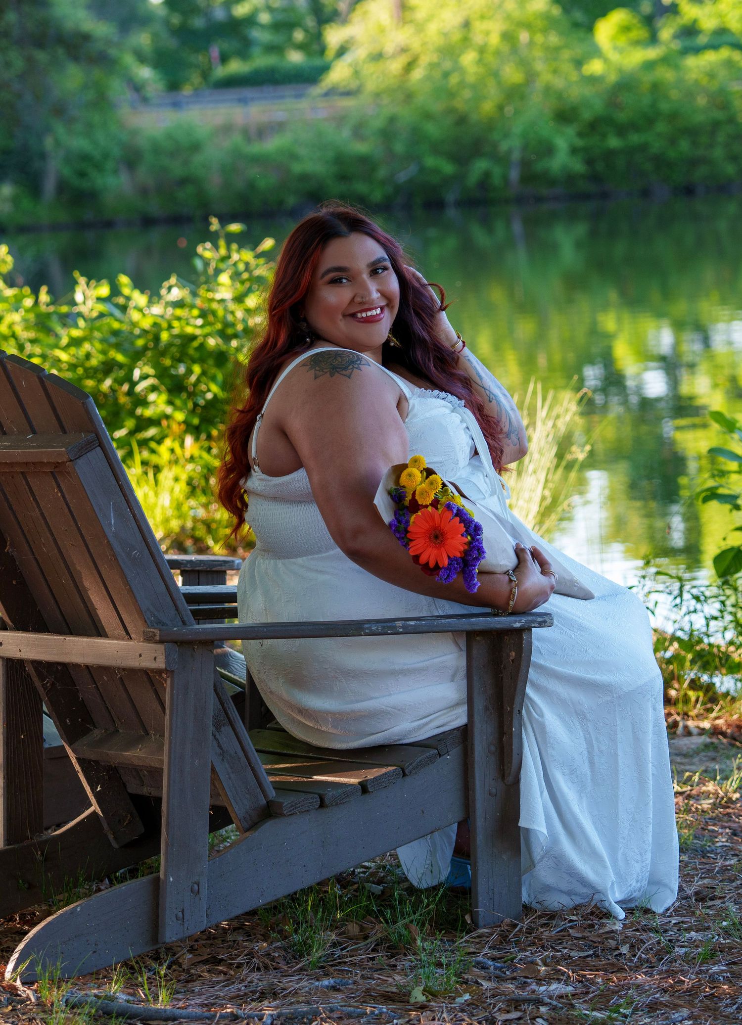 Woman in white dress holding colorful flowers sits in wooden chair by a serene pond surrounded by lush greenery.