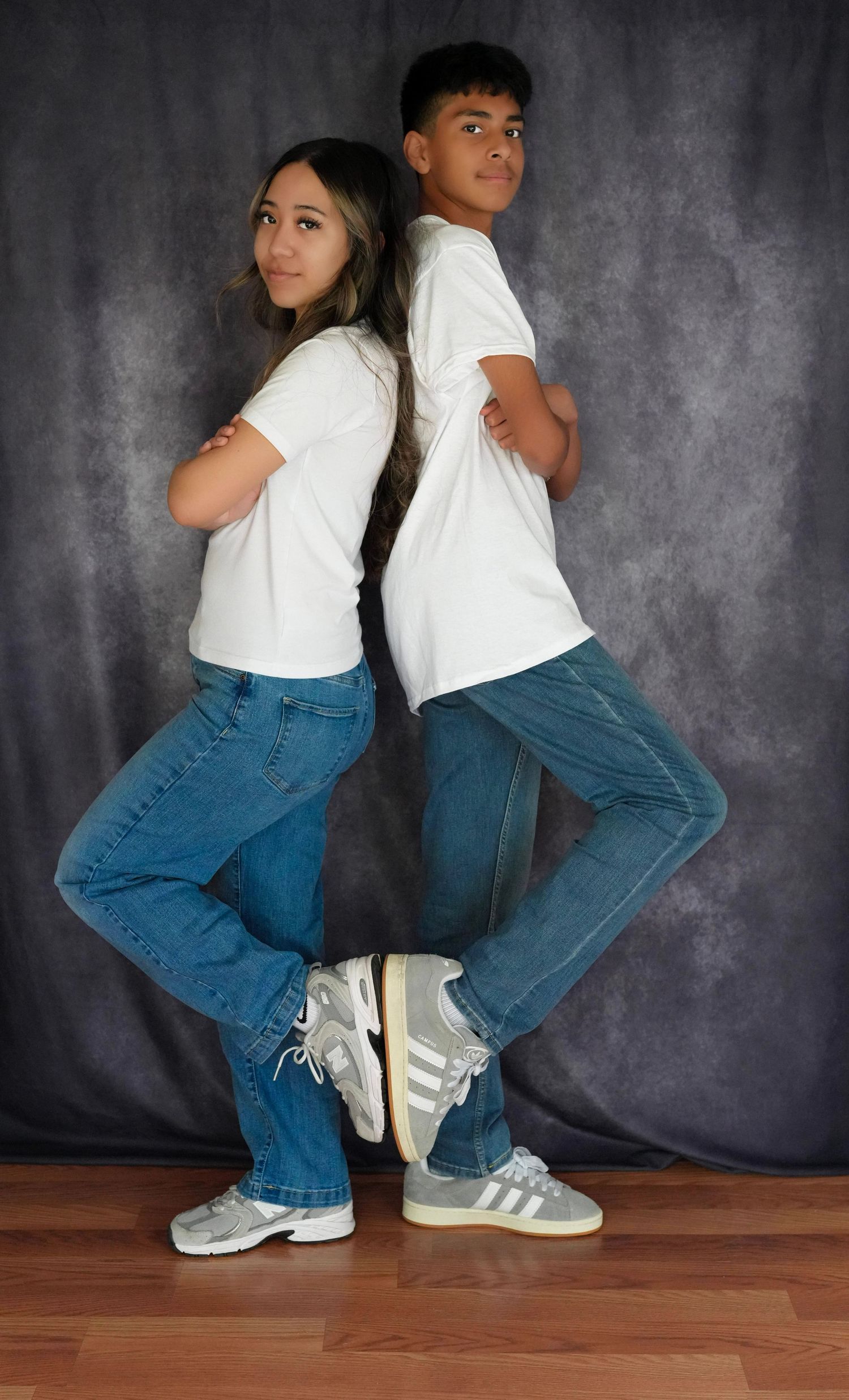 Two teens in white tees and jeans pose back-to-back with arms crossed on a hardwood floor.