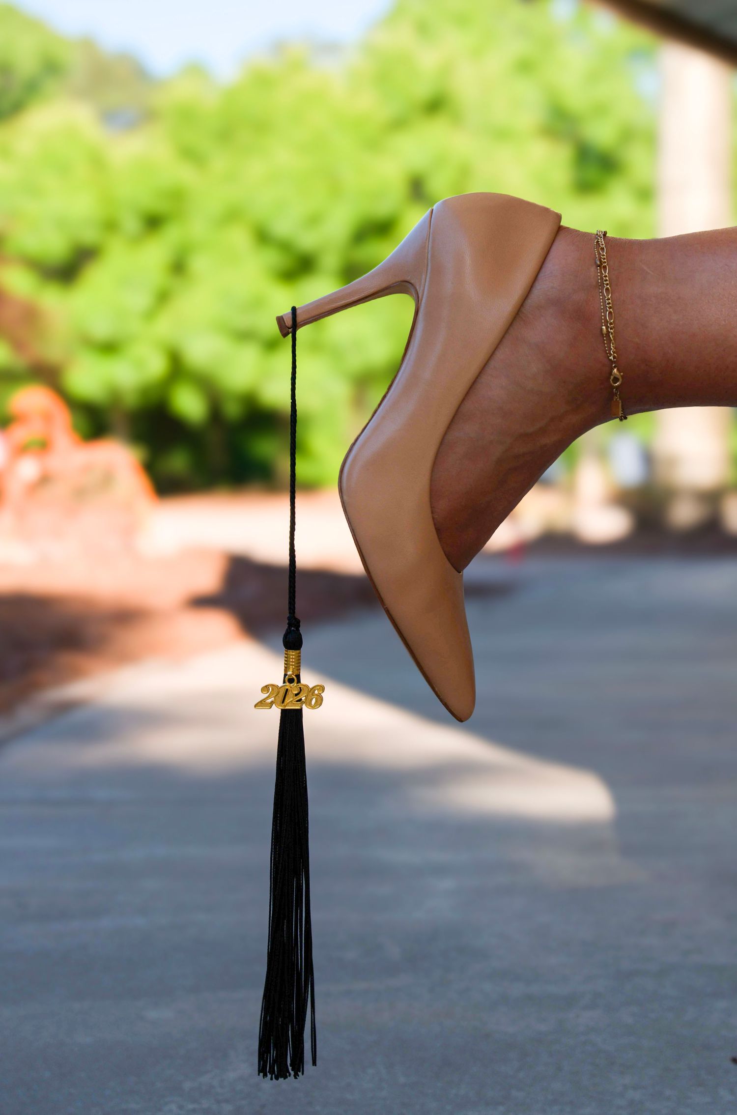 A nude stiletto heel balancing a black graduation tassel with a gold 2026 charm outdoors.