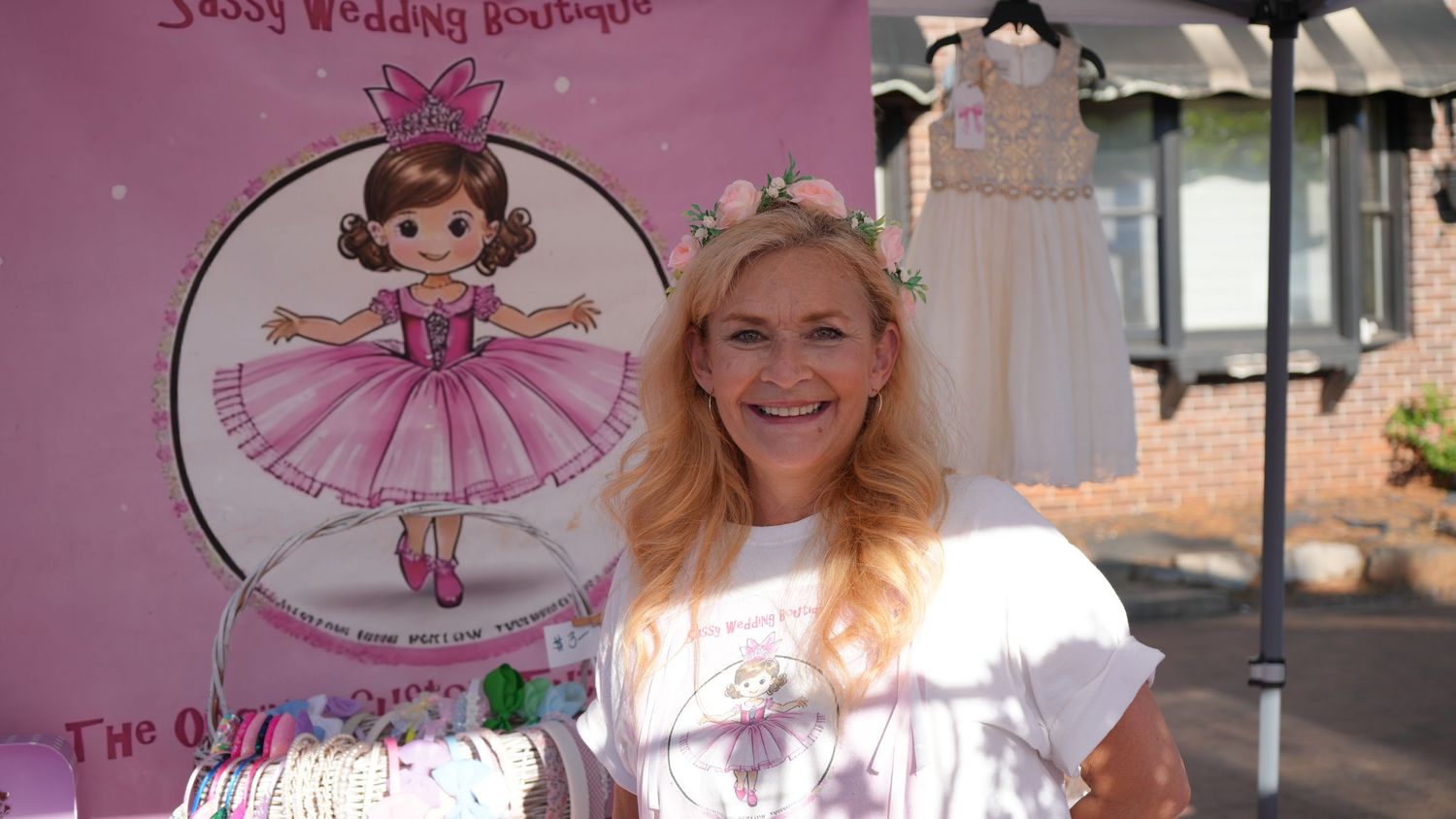 Woman smiling at Sassy Wedding Boutique outdoor vendor booth with children's dresses on display.