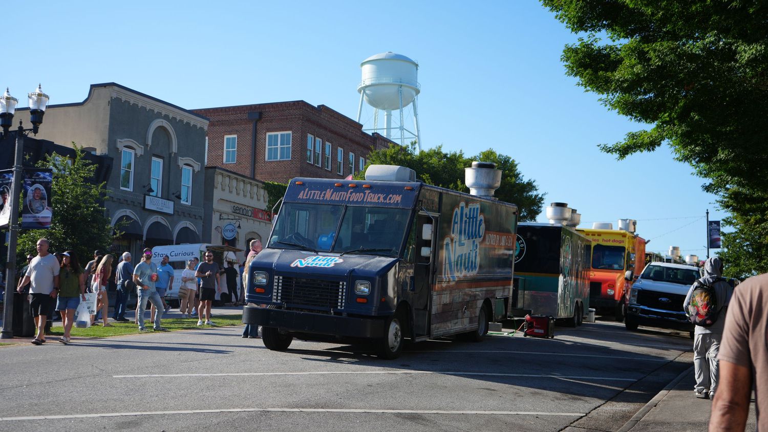 Food trucks lined up on a sunny street in a small town with brick buildings and a water tower in the background.