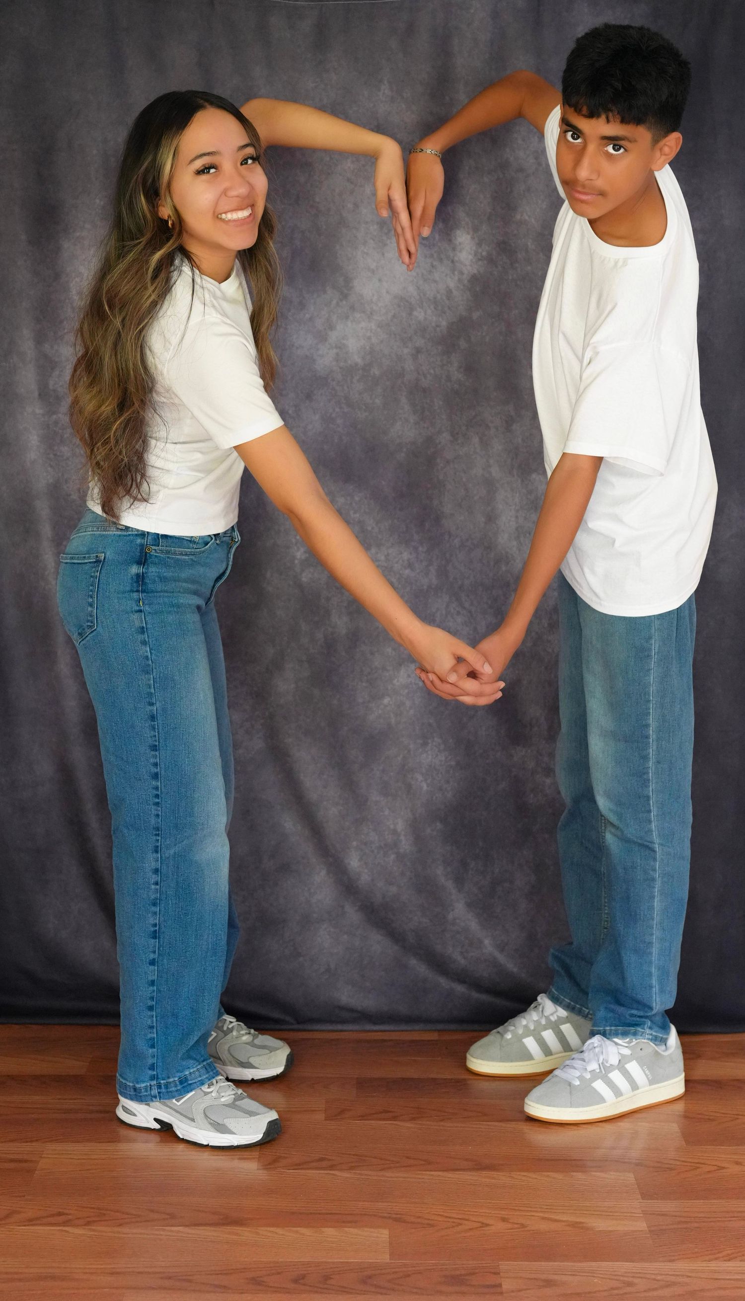 Two teens in matching white tees and jeans form a heart shape with their arms against a gray backdrop.
