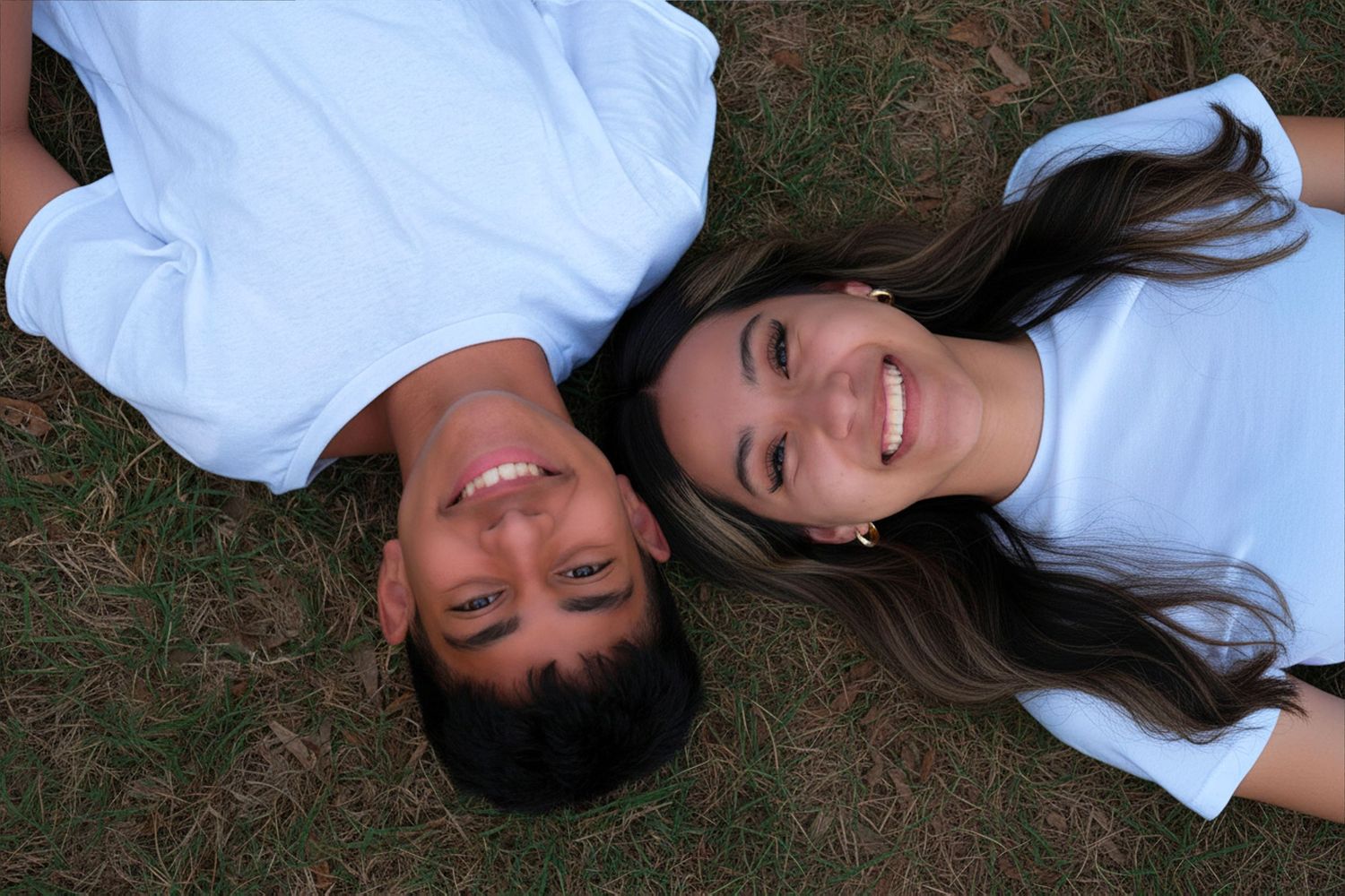 Two smiling young people lying on green grass, wearing white t-shirts, looking up at the camera.
