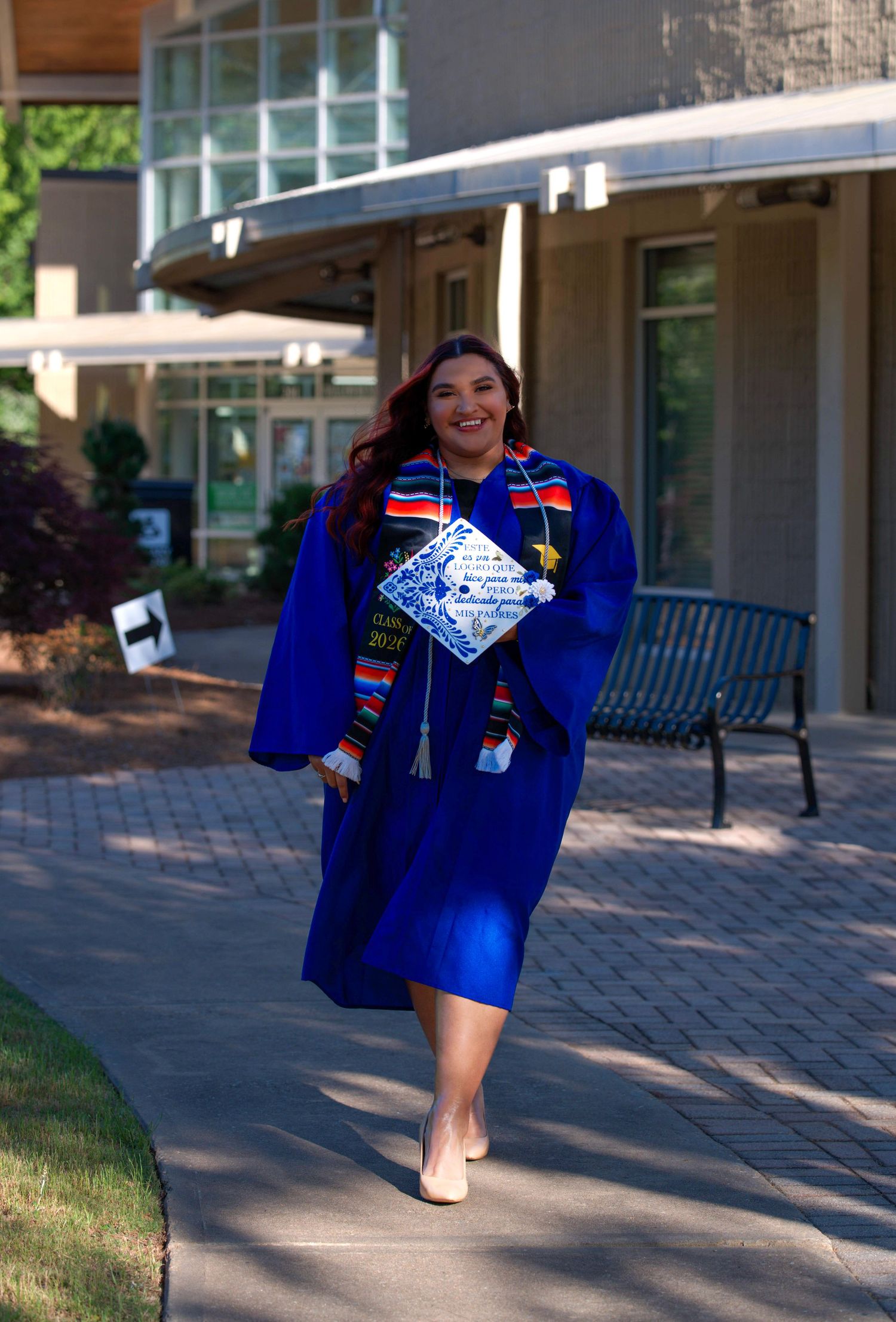 Smiling woman in blue graduation gown and colorful stole walks on campus pathway.