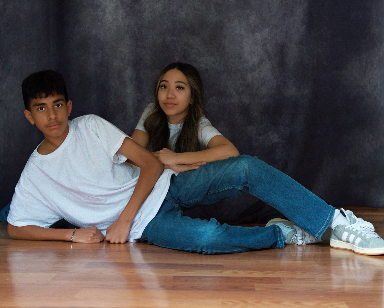 Two teenagers pose on a wooden floor against a dark gray backdrop, wearing white t-shirts and jeans.