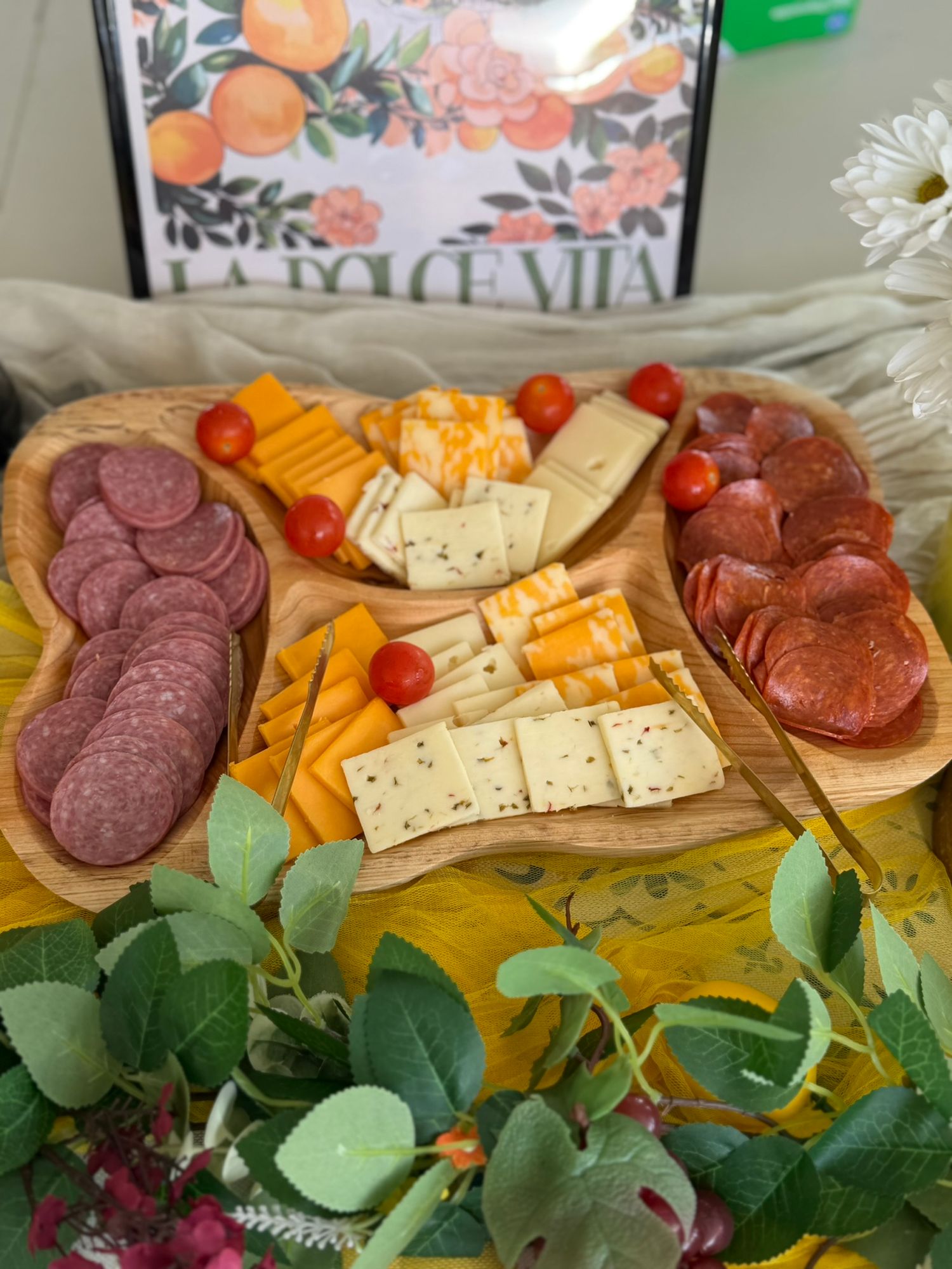 Wooden charcuterie board with salami, cheddar cheese, crackers, and cherry tomatoes on a floral table setting.