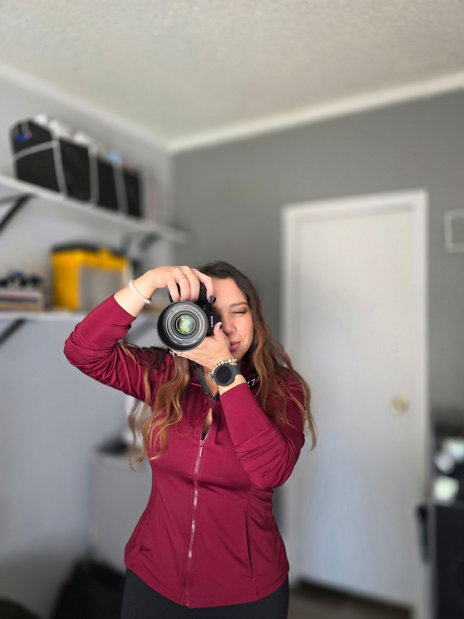 Photographer in burgundy sweater demonstrates different camera angles and poses while taking photos in a bright room.
