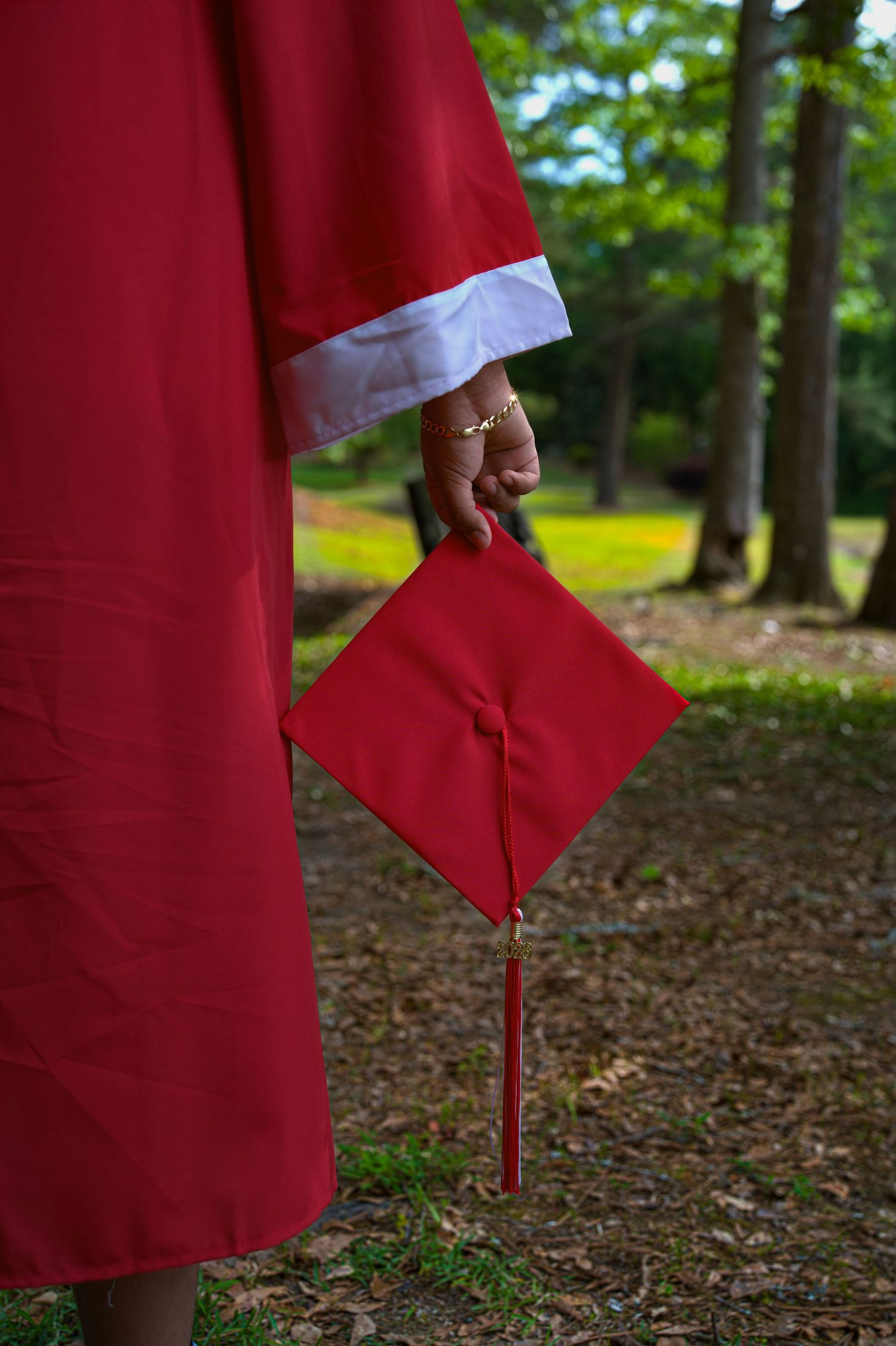 Graduate in red cap and gown holding mortarboard with tassel in a lush green outdoor setting.