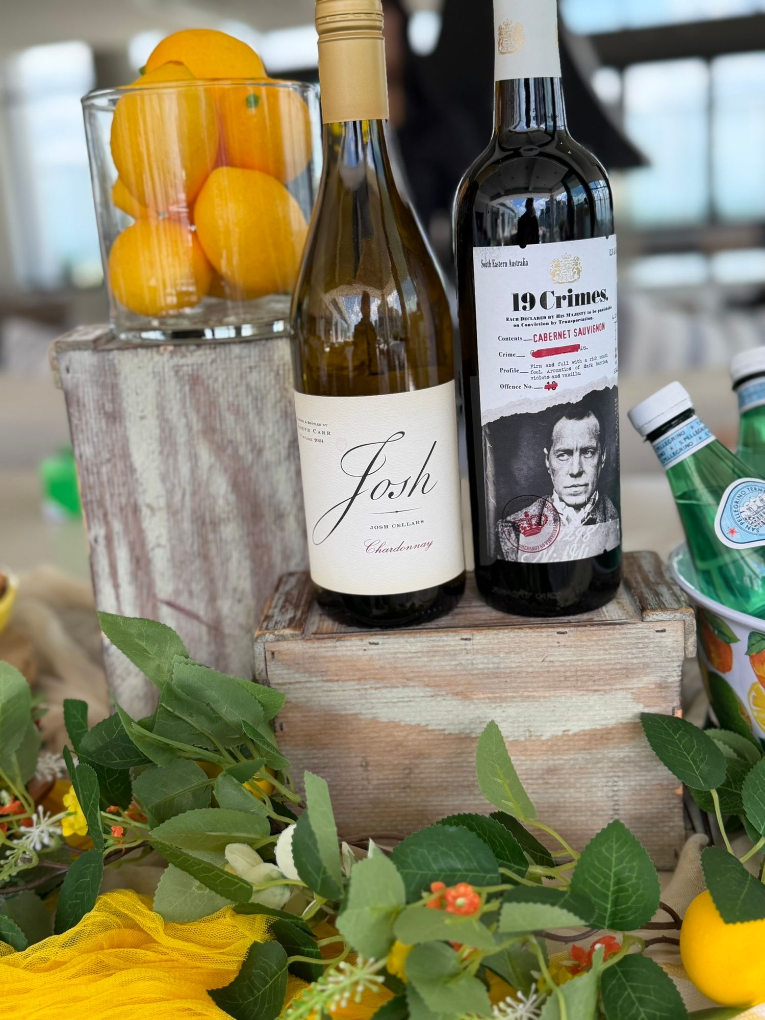 Two wine bottles, Josh Chardonnay and 19 Crimes Cabernet Sauvignon, displayed on a wooden box with floral decor.