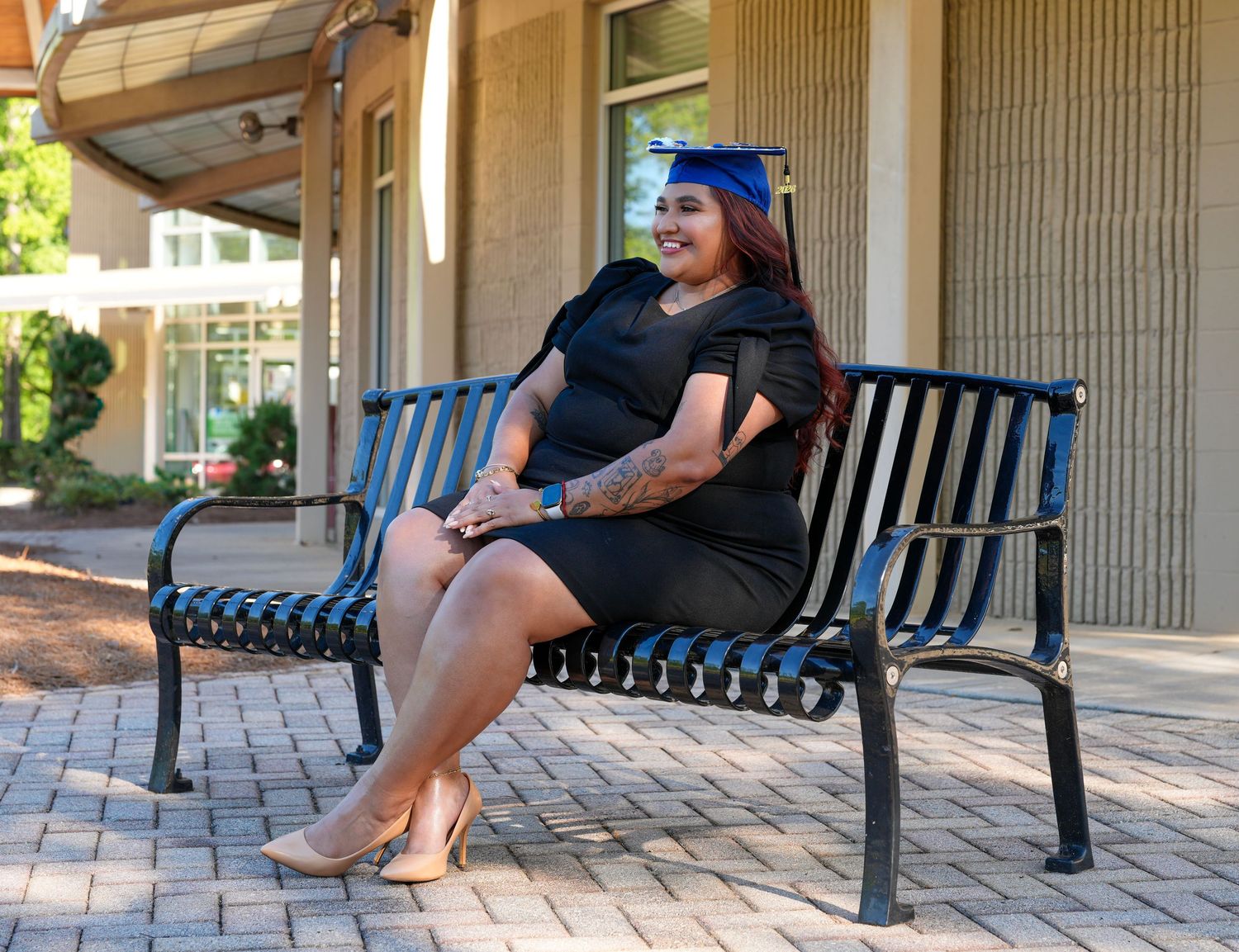 Woman in black dress and graduation cap smiling on outdoor bench, celebrating academic achievement.