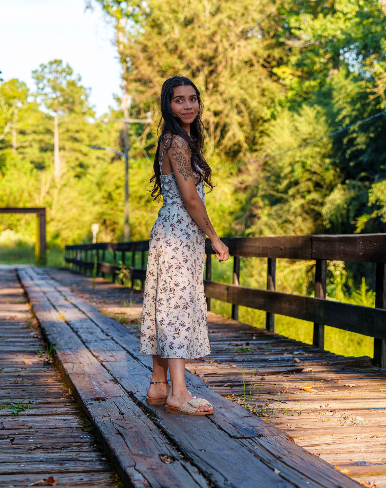 Young woman in floral midi dress and sandals posing on a wooden bridge surrounded by lush green trees.