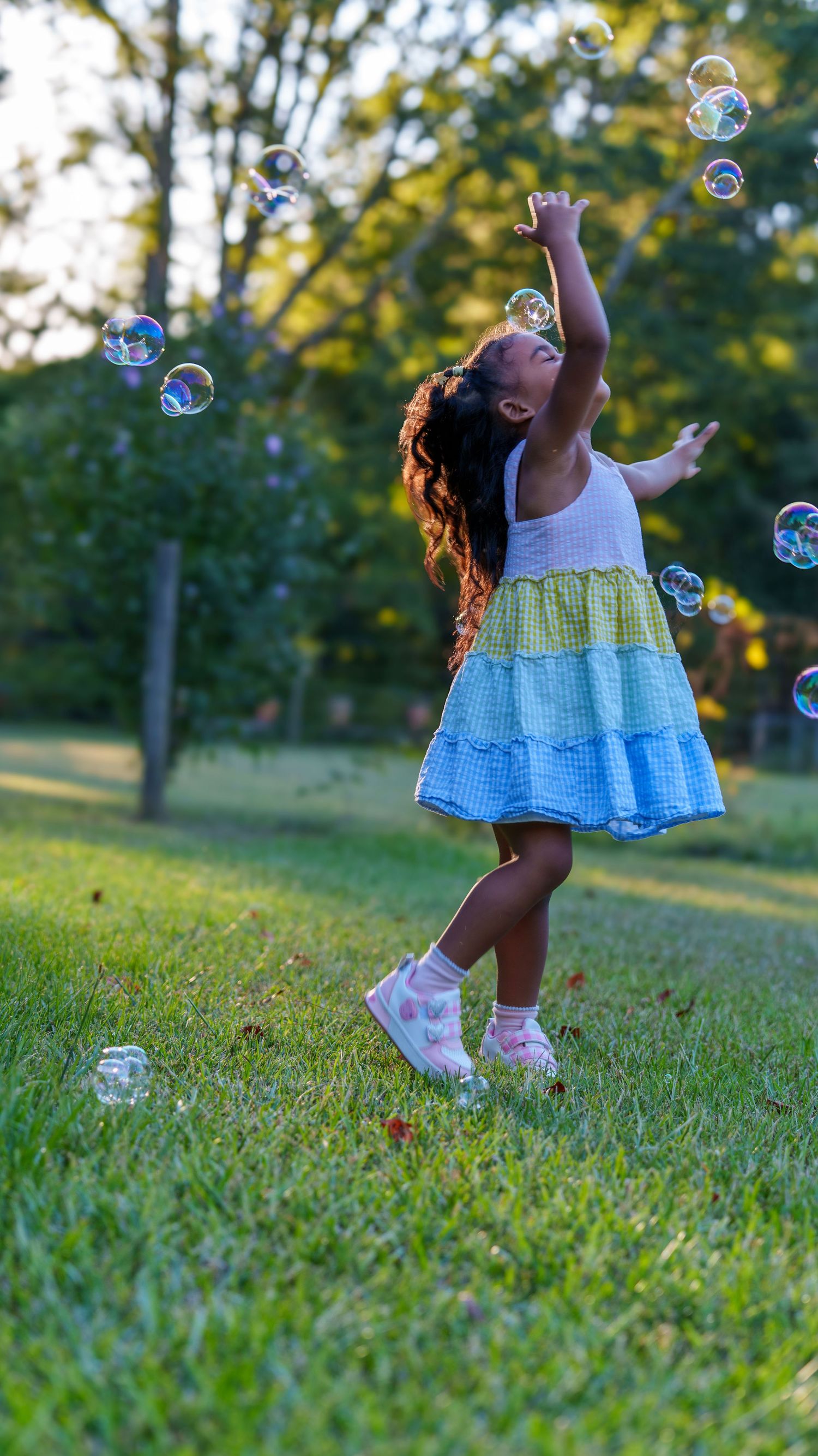 A young girl in a colorful tiered dress joyfully reaches for soap bubbles floating in a sunny green park.