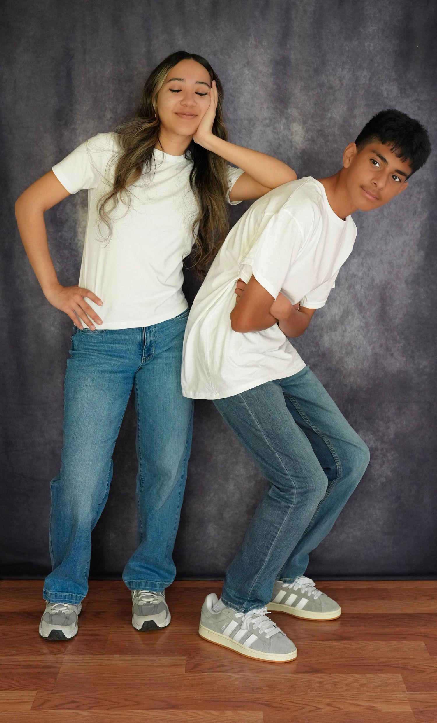 Two teens in white t-shirts and blue jeans pose confidently against a gray backdrop on a wood floor.