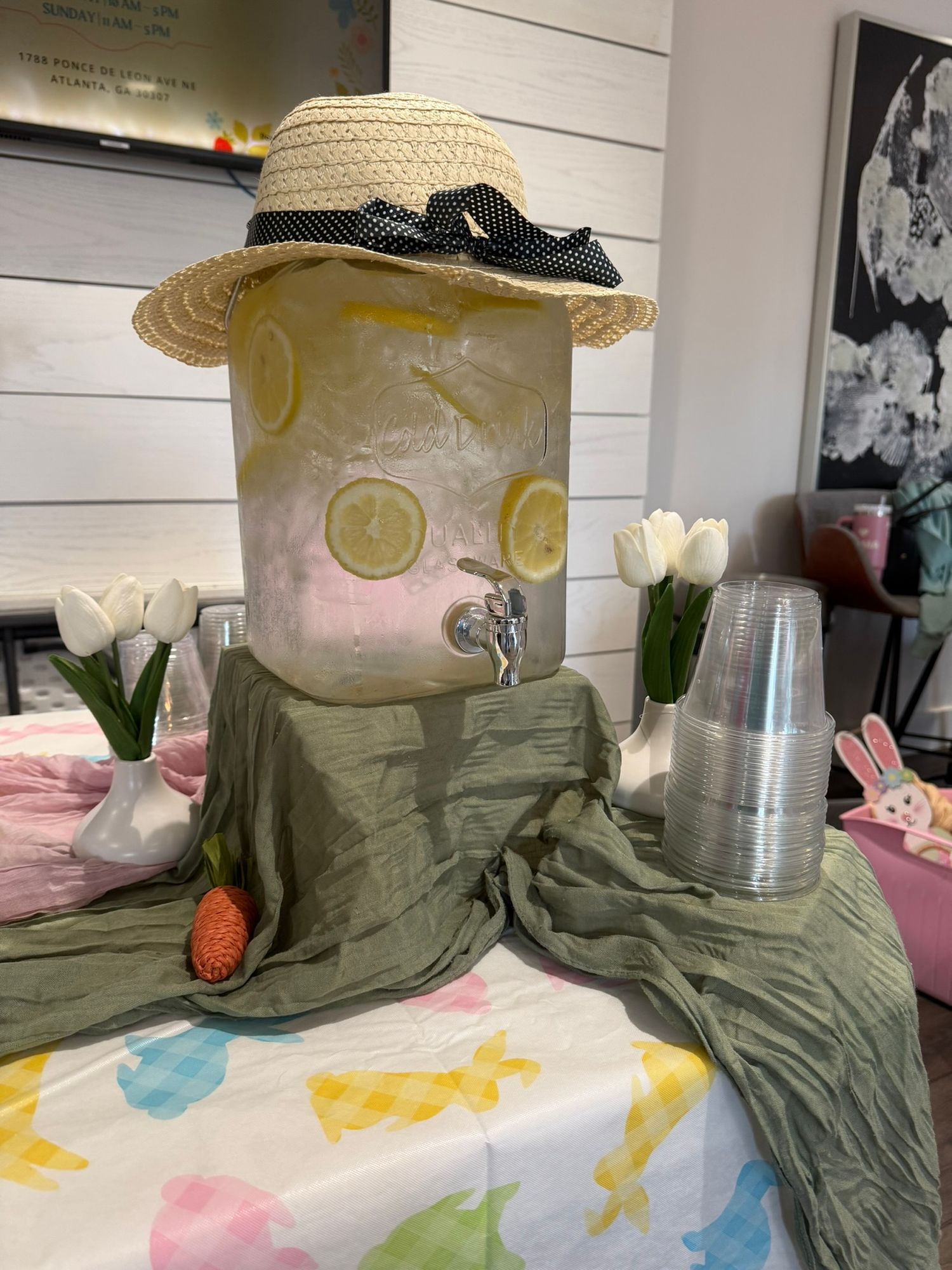 A lemonade drink dispenser decorated with a straw hat, lemon slices, and white tulips on a festive Easter-themed table.