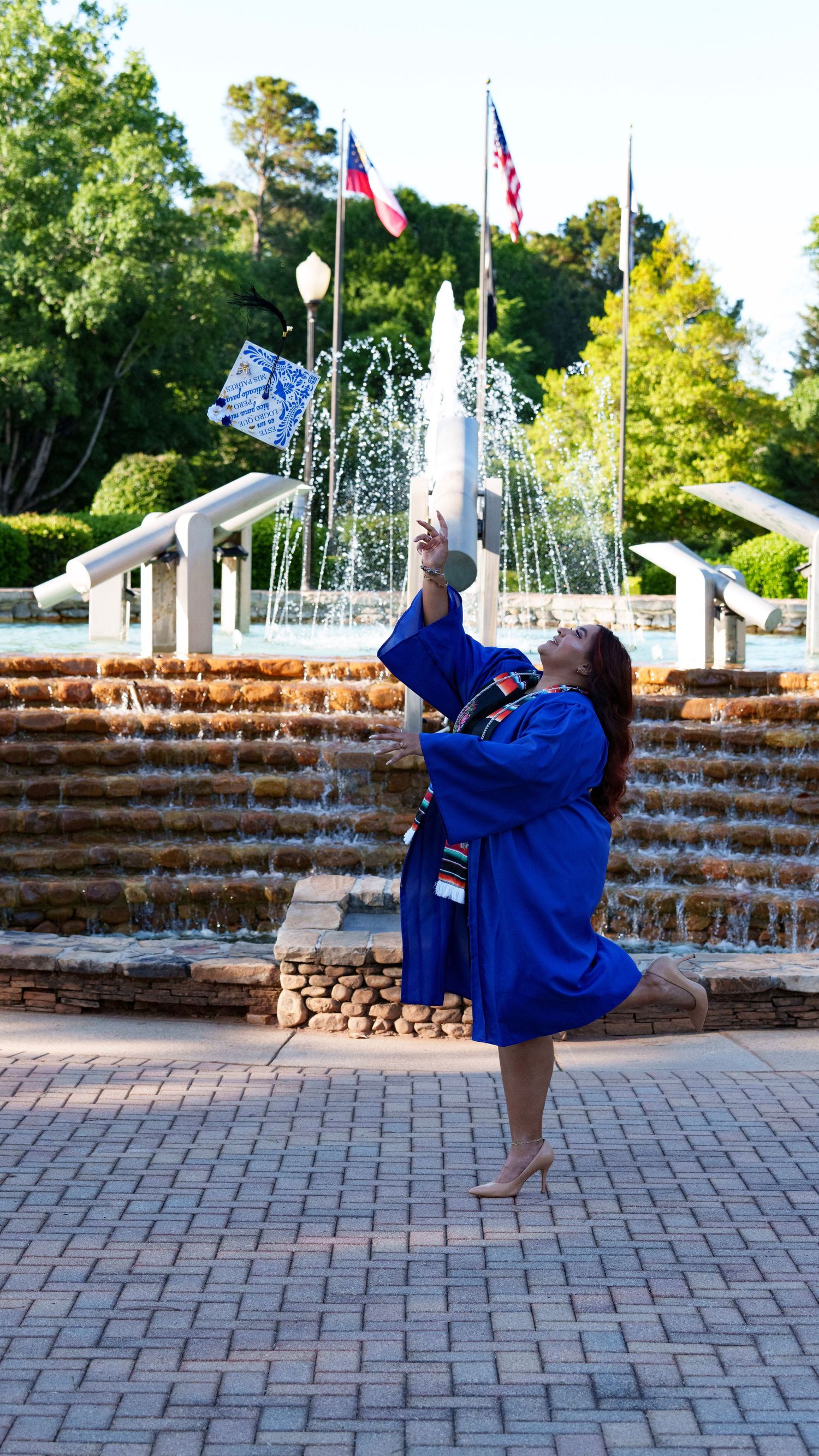 Graduate in blue gown tossing cap near fountain with flags, celebrating graduation outdoors on brick plaza.