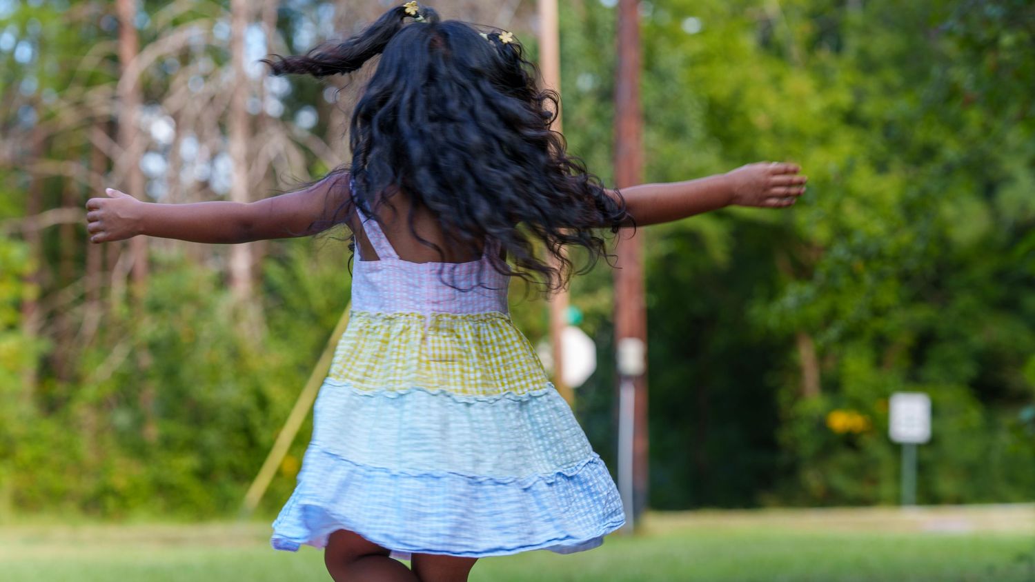 Young Black girl in colorful tiered dress spinning outdoors with arms outstretched, hair flying, in a green park setting.
