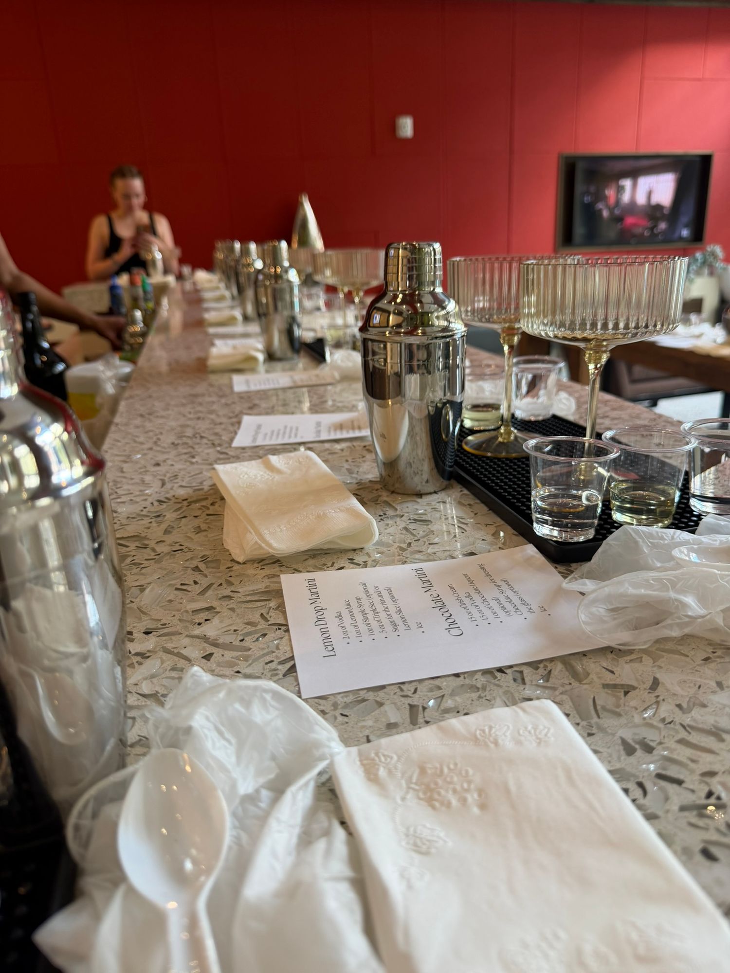 Long marble bar setup for cocktail making class with shakers, glassware, napkins, and recipe cards at each station.