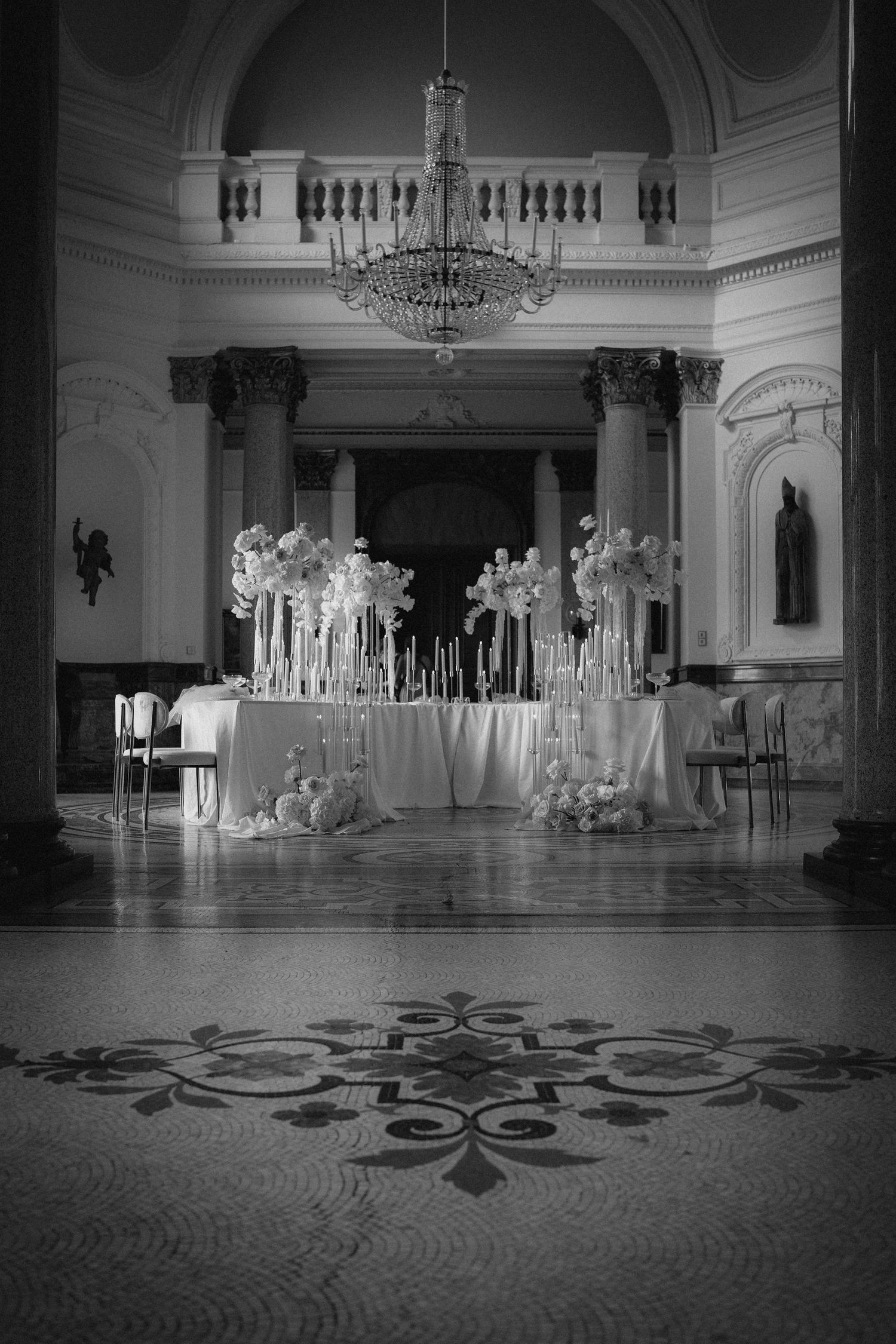 Elegant black and white interior with crystal chandelier and floral arrangements on display table in grand ballroom setting.