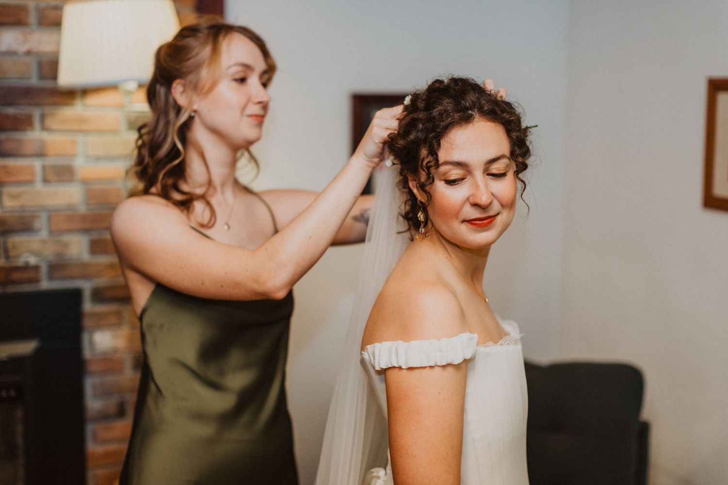 Bridesmaid in olive dress helping bride with veil during wedding day preparations indoors.