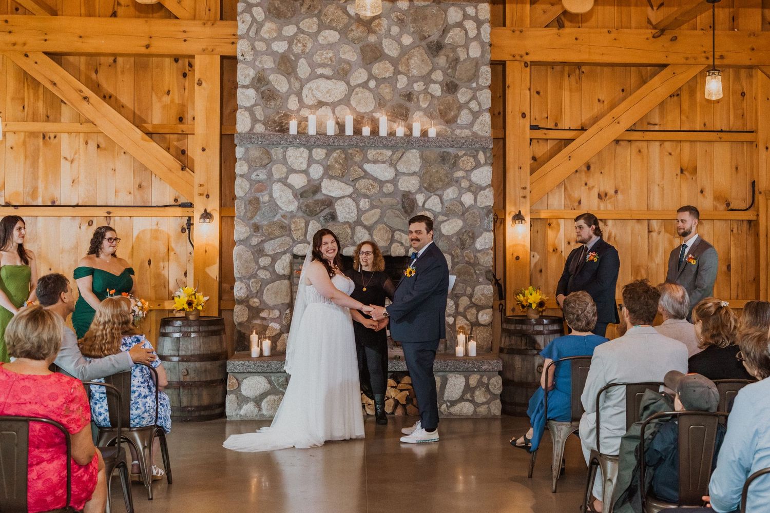 Bride and groom exchange vows at rustic barn wedding ceremony with stone fireplace backdrop and wooden decor.