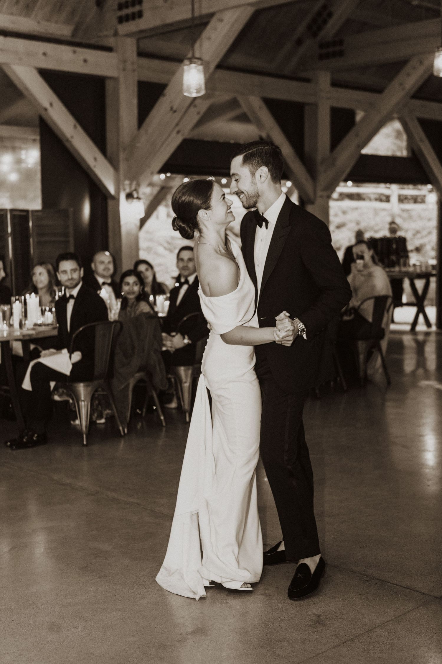Black and white photo of bride and groom sharing their first dance in a rustic wooden venue.