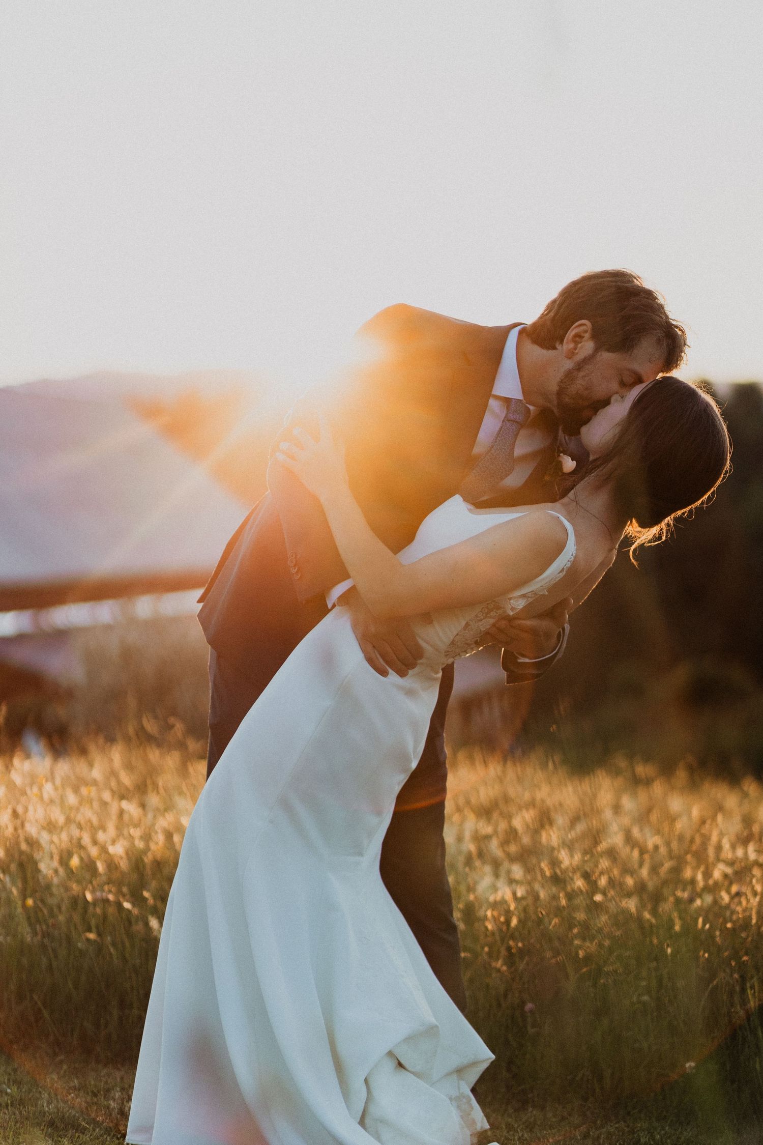 Bride and groom sharing romantic dip kiss at golden hour sunset in an open field at wedding.
