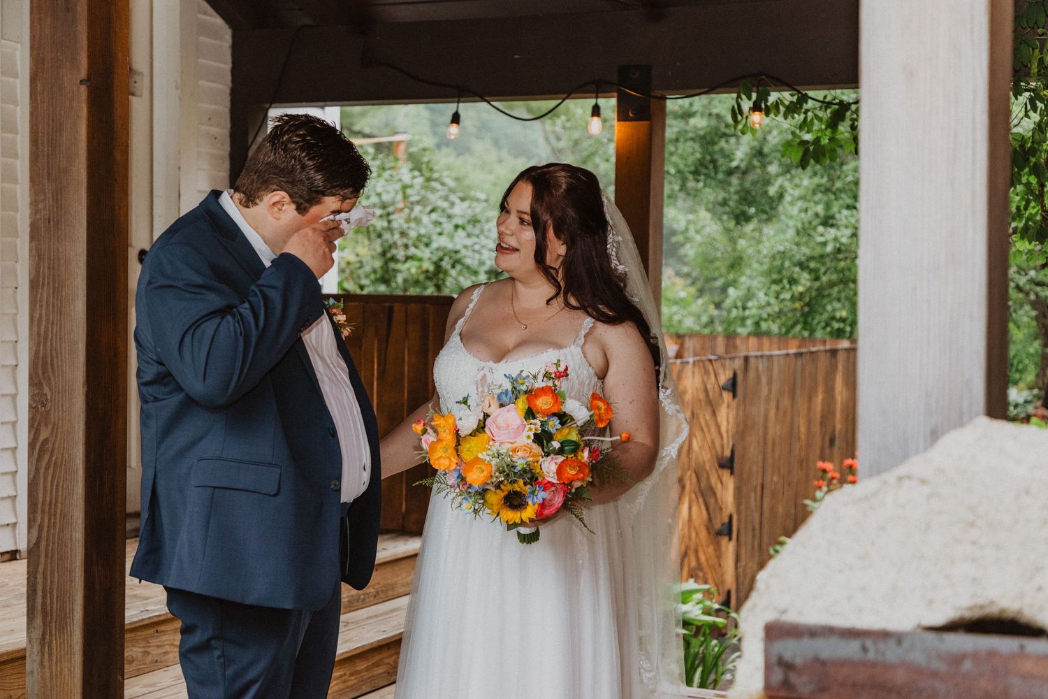Groom wipes tears seeing bride in white gown holding sunflower bouquet during outdoor wedding first look.