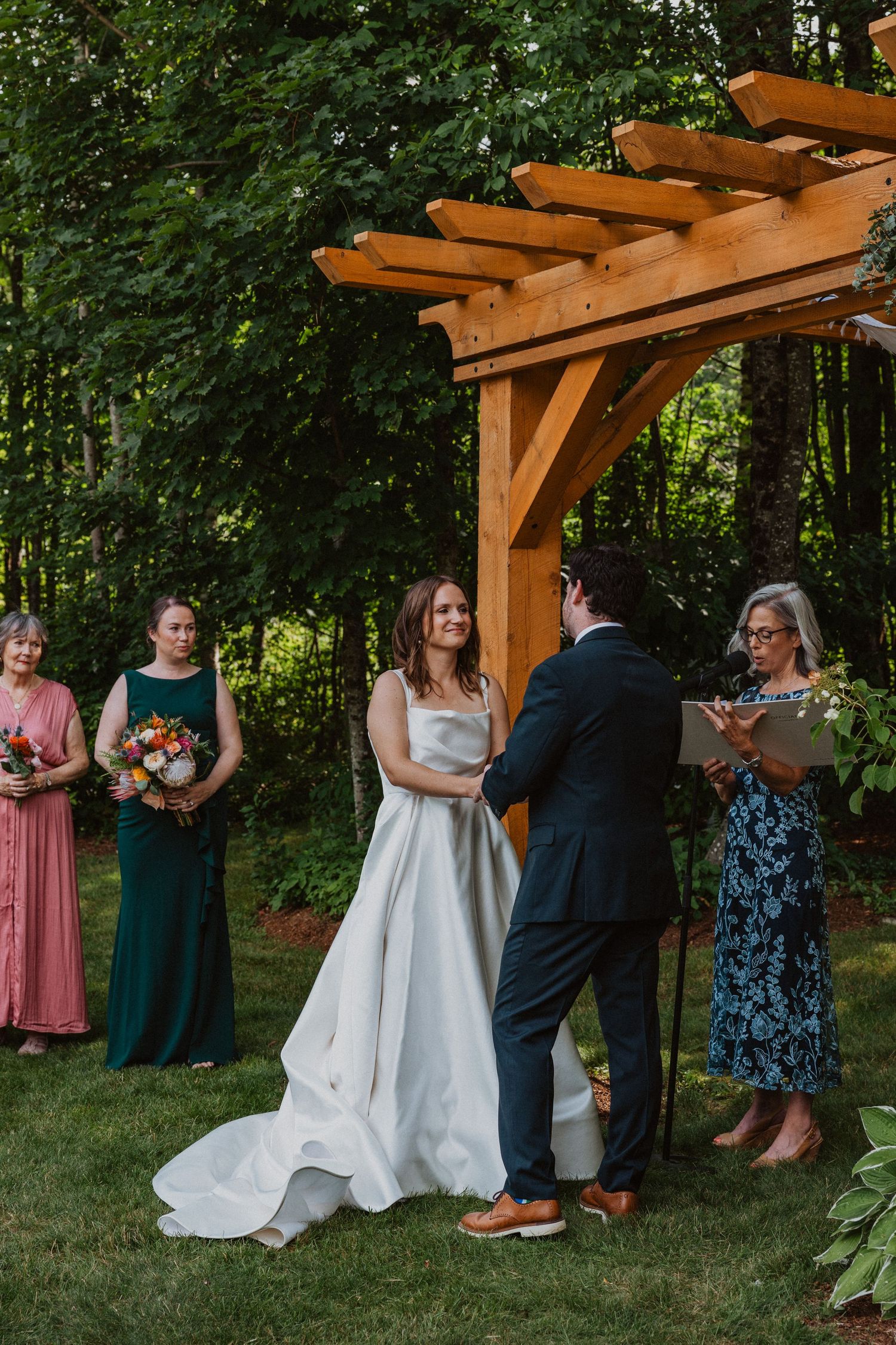 Bride and groom exchange vows under wooden pergola with bridesmaids in pink and green dresses beside them.
