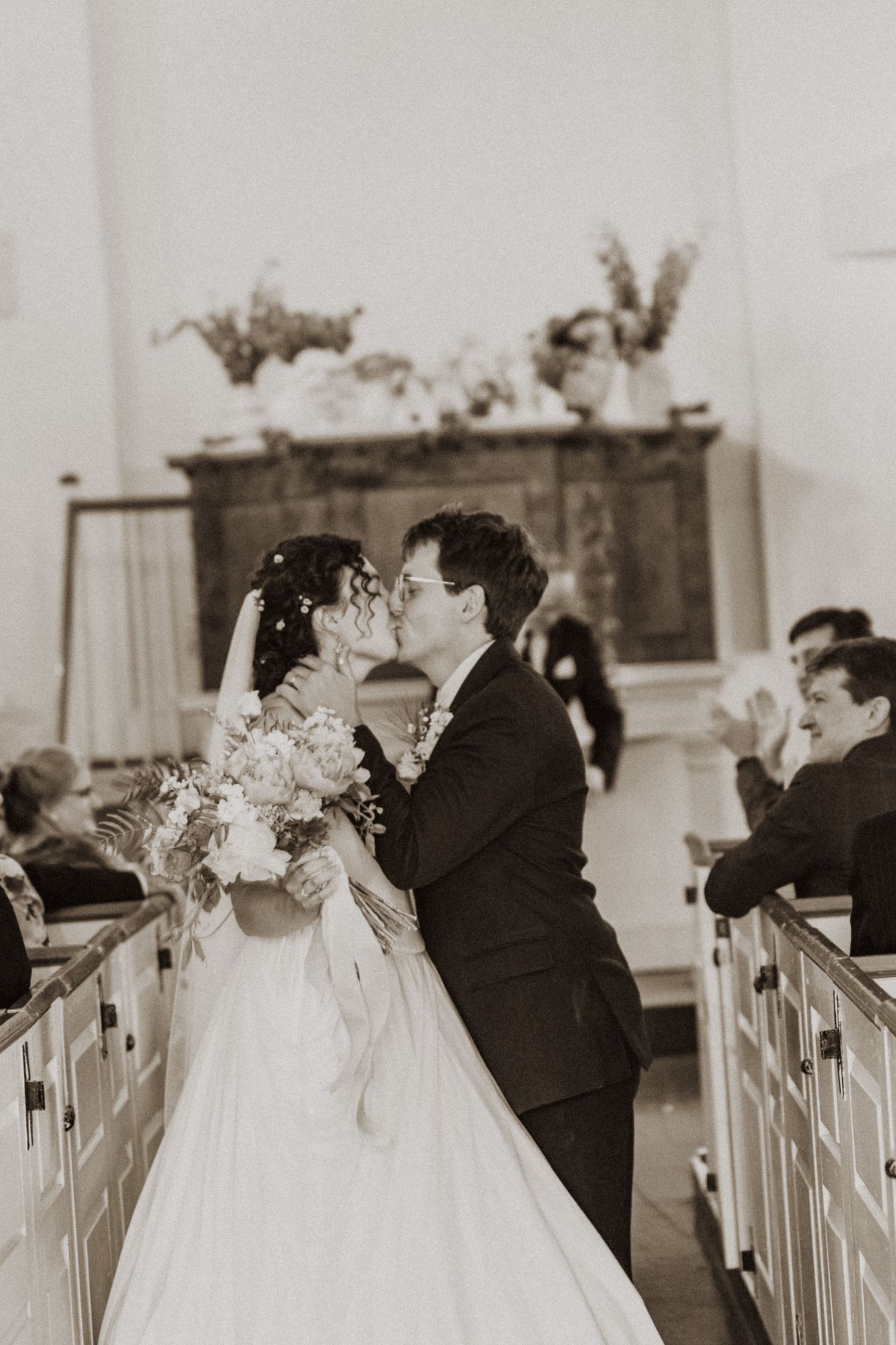 Sepia toned bride and groom sharing first kiss at church altar surrounded by wedding guests.