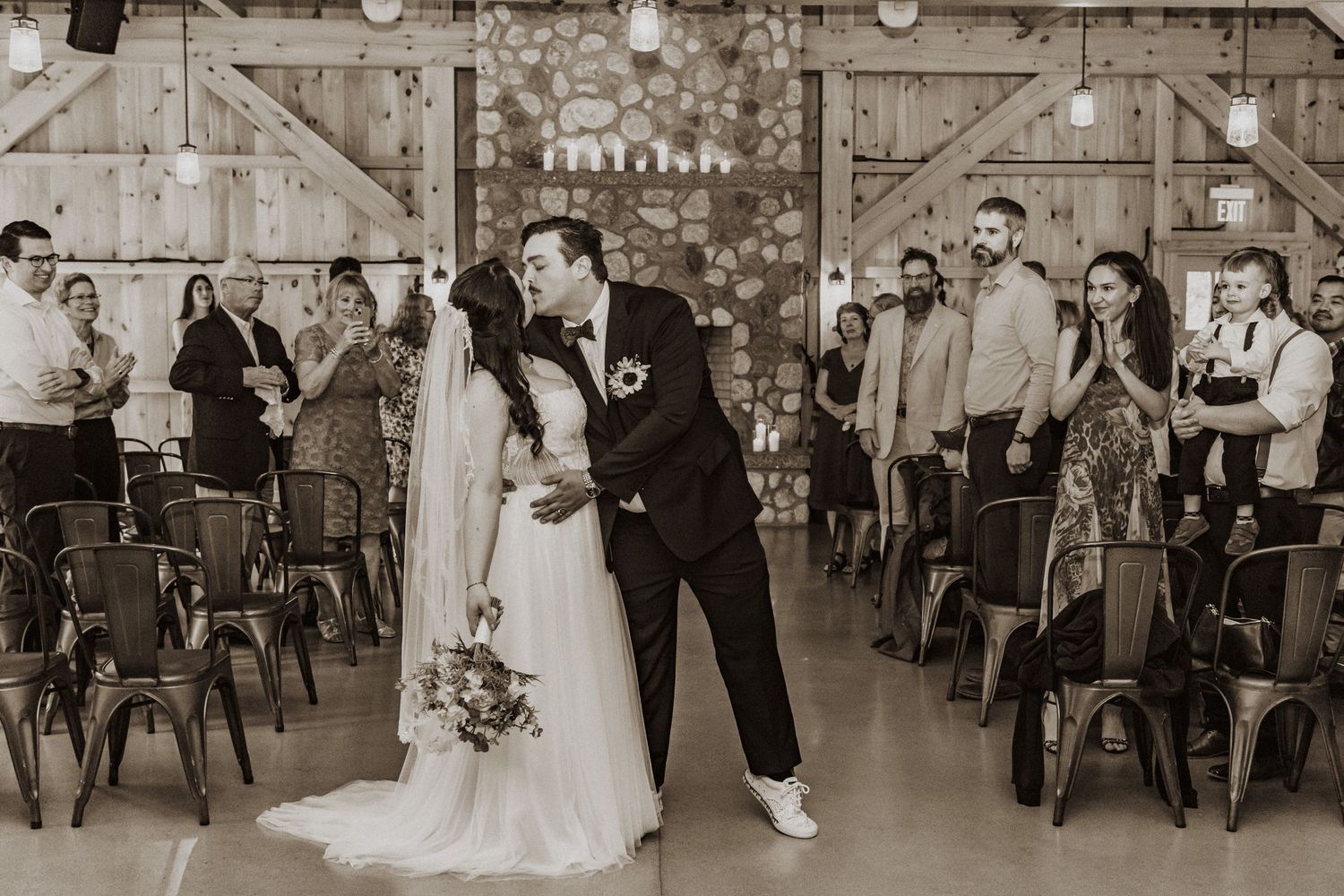 Bride and groom share first kiss as newlyweds walking down aisle in black and white photo.