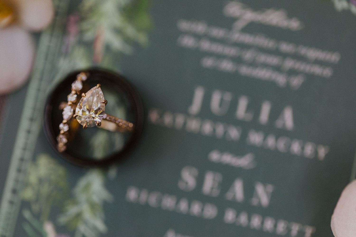 Elegant marquise diamond engagement ring beside a green wedding invitation for Julia and Sean on a dark ring box.