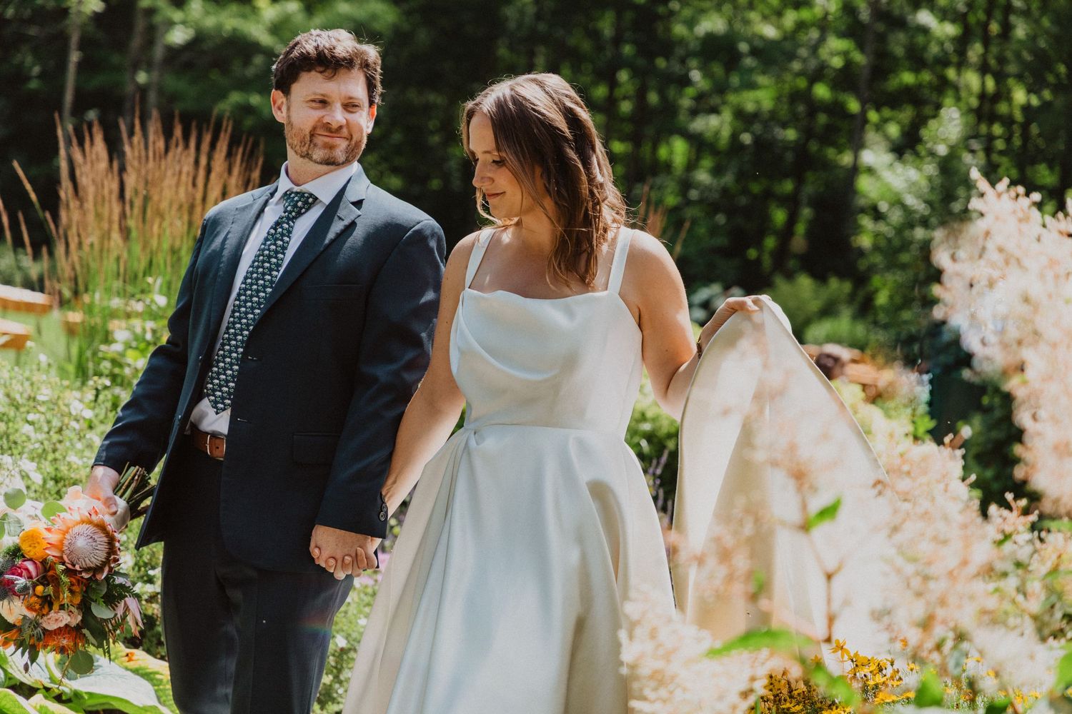 Bride and groom holding hands smiling outdoors, surrounded by lush greenery and colorful floral arrangements.